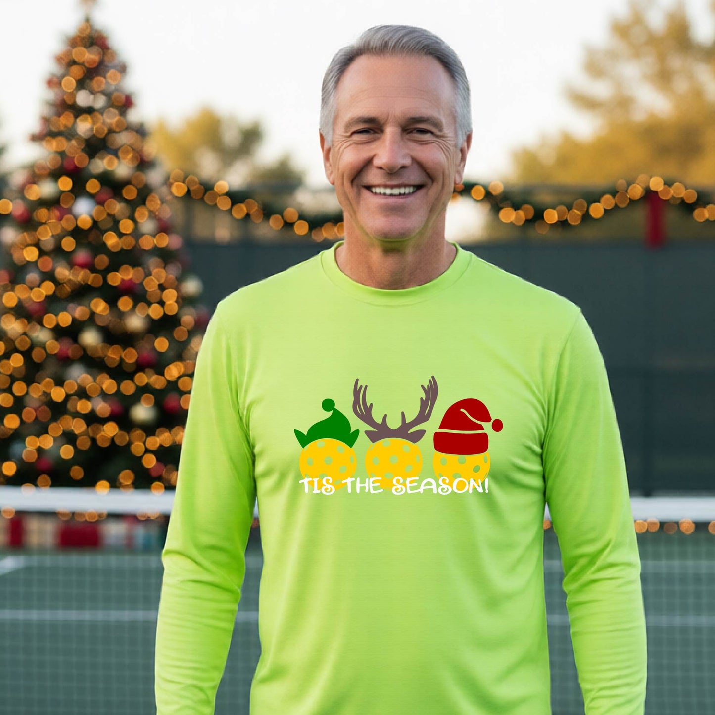 A man wearing a lime green graphic long-sleeve t-shirt with a Christmas-themed design of three pickleballs wearing holiday hats and the text 'TIS THE SEASON!'. The background shows a festive blurred Christmas tree and a pickleball court.