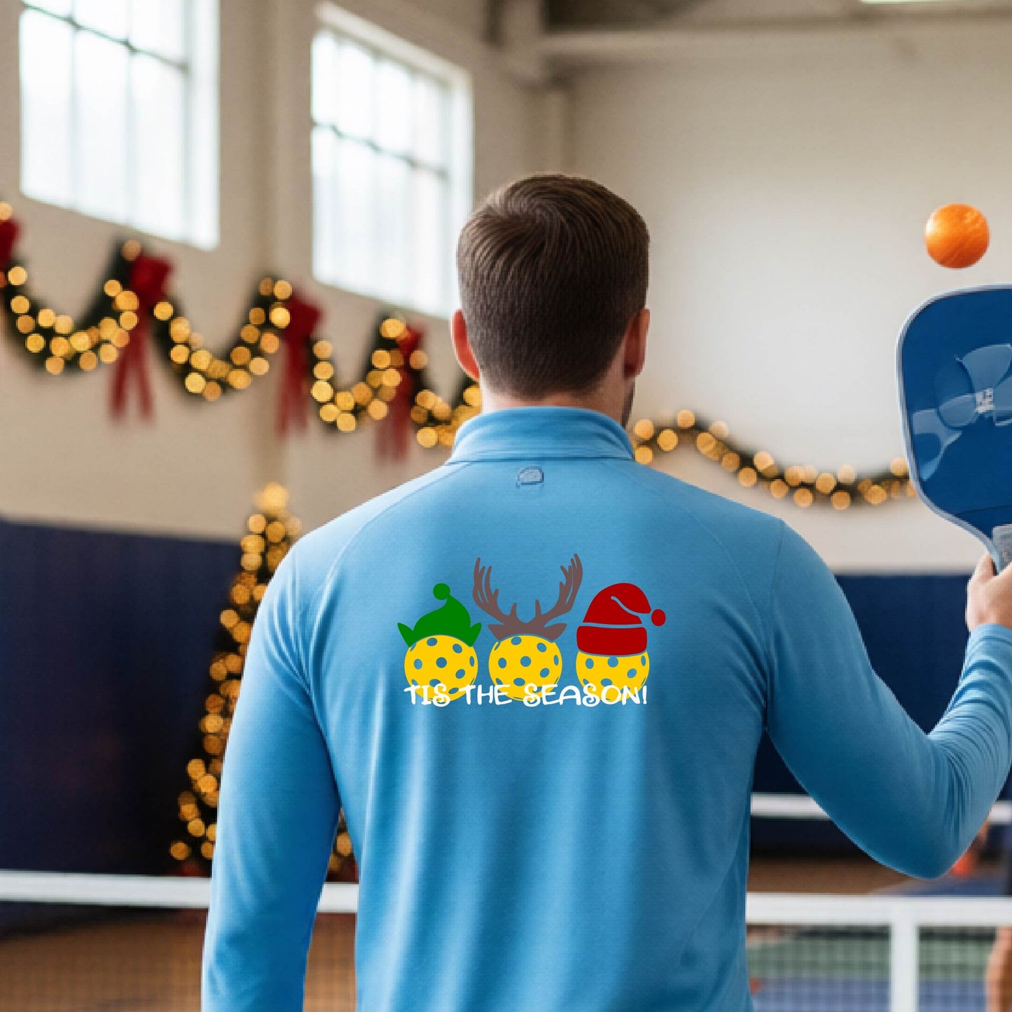 An man viewed from behind in a gymnasium, wearing a light blue long-sleeve quarter-zip shirt with a Christmas-themed back graphic showing three decorated pickleball and the text 'TIS THE SEASON!'.
