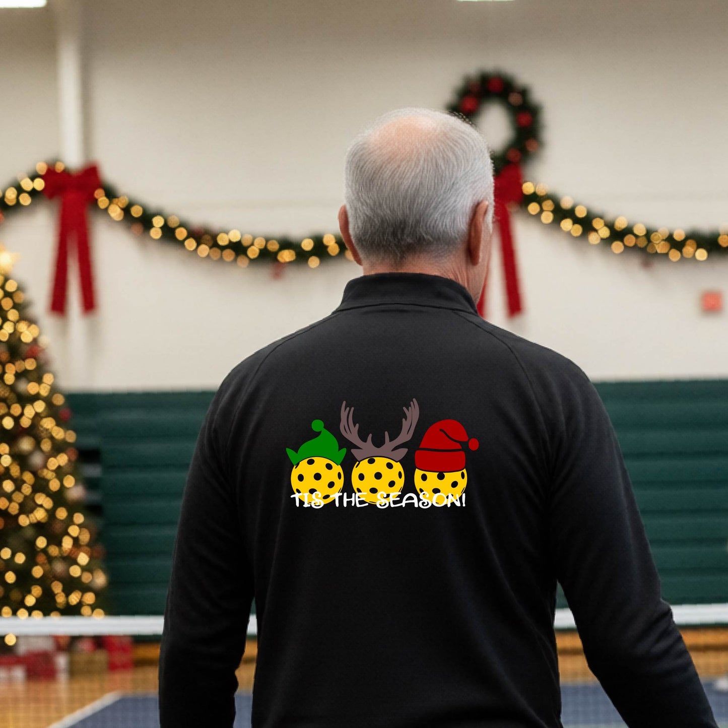 An man viewed from behind in a gymnasium, wearing a black long-sleeve quarter-zip shirt with a Christmas-themed back graphic showing three decorated pickleball and the text 'TIS THE SEASON!'.