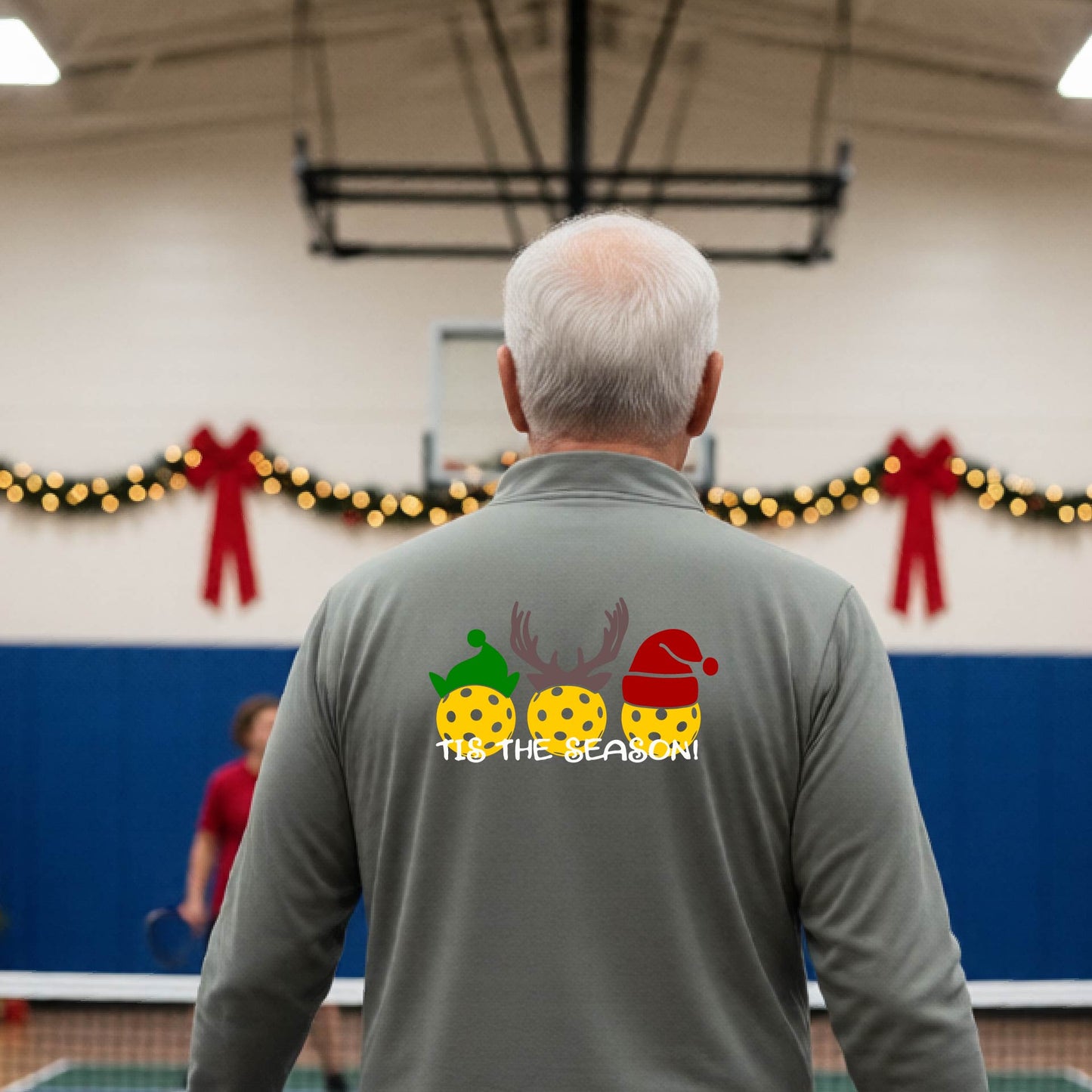 An man viewed from behind in a gymnasium, wearing a gray long-sleeve quarter-zip shirt with a Christmas-themed back graphic showing three decorated pickleball and the text 'TIS THE SEASON!'.