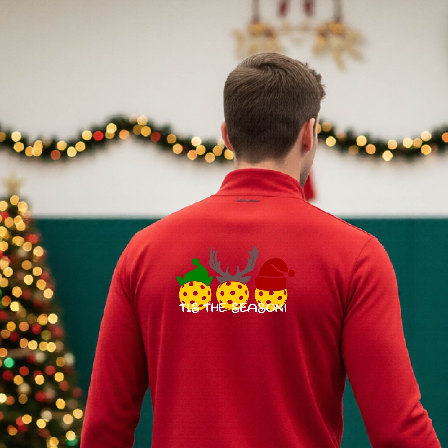 An man viewed from behind in a gymnasium, wearing a red long-sleeve quarter-zip shirt with a Christmas-themed back graphic showing three decorated pickleball and the text 'TIS THE SEASON!'.