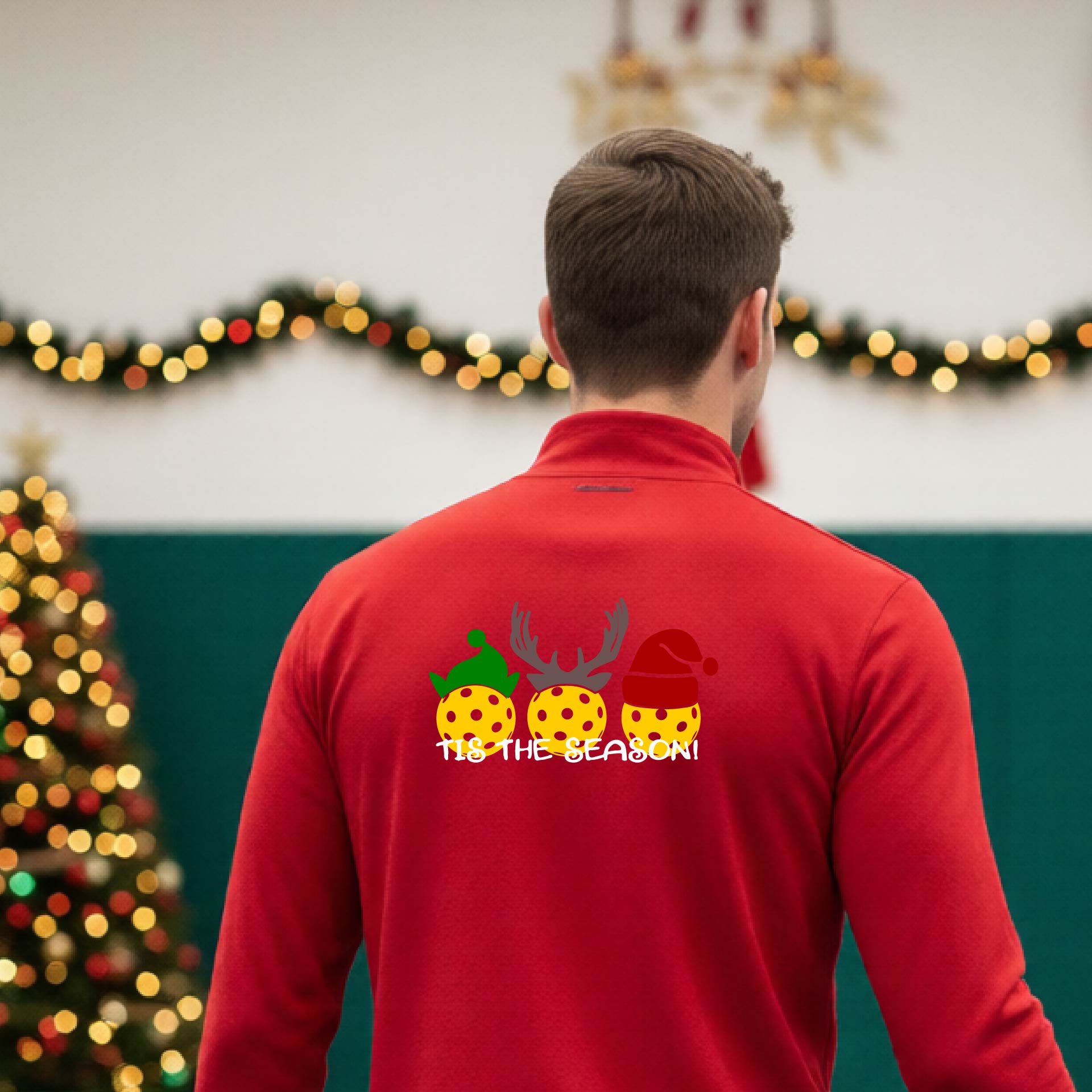 An man viewed from behind in a gymnasium, wearing a red long-sleeve quarter-zip shirt with a Christmas-themed back graphic showing three decorated pickleball and the text 'TIS THE SEASON!'.