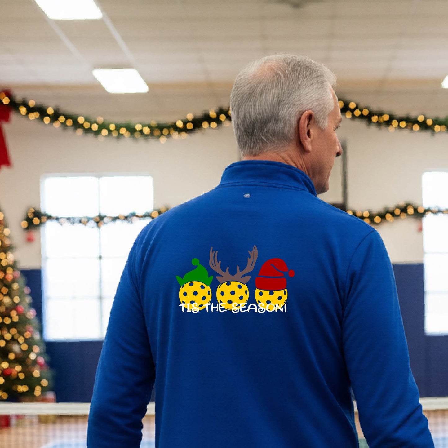 An man viewed from behind in a gymnasium, wearing a royal blue long-sleeve quarter-zip shirt with a Christmas-themed back graphic showing three decorated pickleball and the text 'TIS THE SEASON!'.