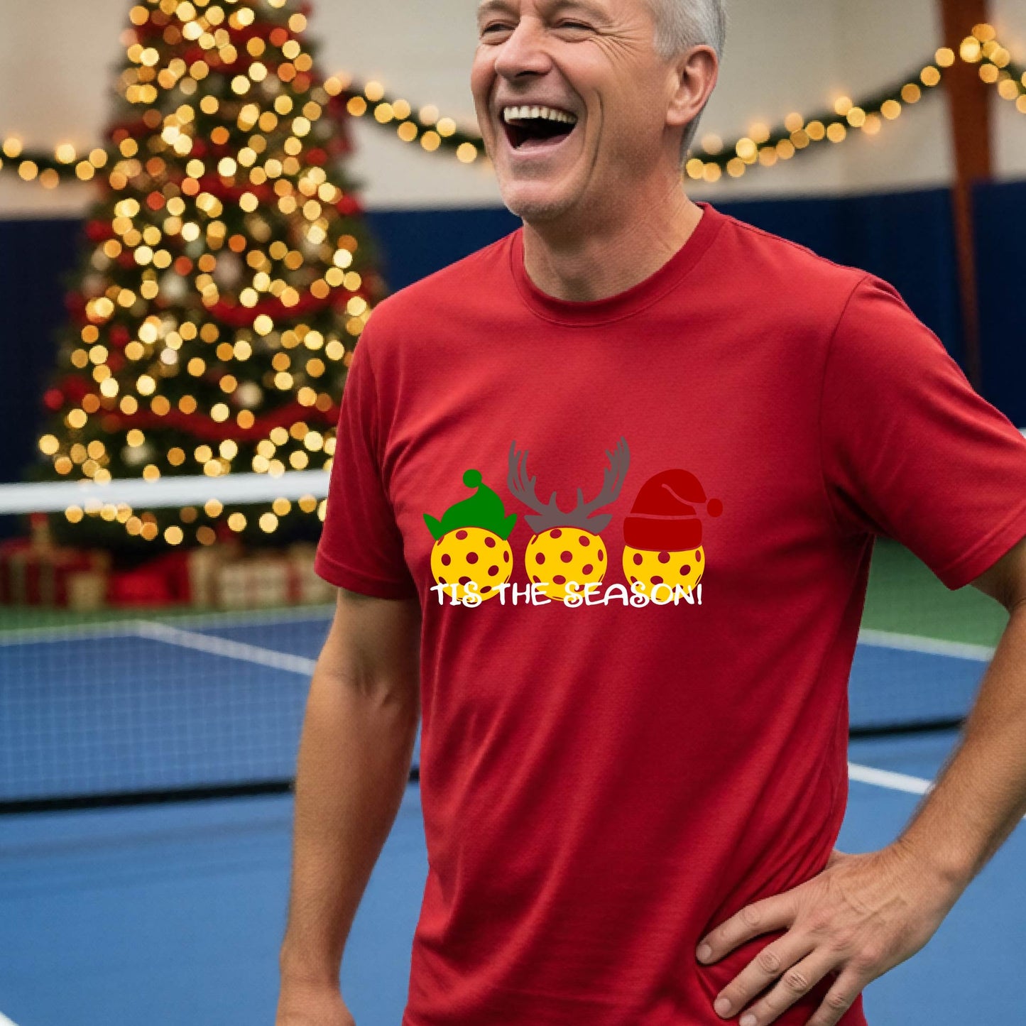 A man wearing a red  graphic short-sleeve t-shirt with a Christmas-themed design of three pickleballs wearing holiday hats and the text 'TIS THE SEASON!'. The background shows a festive blurred Christmas tree and a pickleball court.
