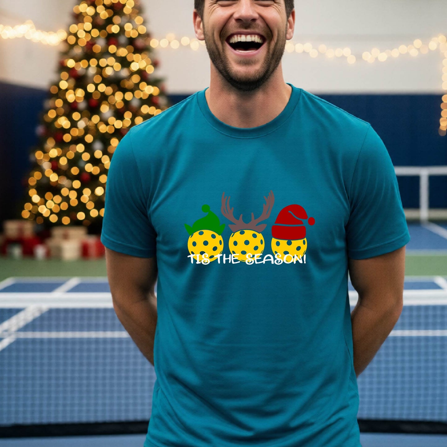 A man wearing a teal graphic short-sleeve t-shirt with a Christmas-themed design of three pickleballs wearing holiday hats and the text 'TIS THE SEASON!'. The background shows a festive blurred Christmas tree and a pickleball court.