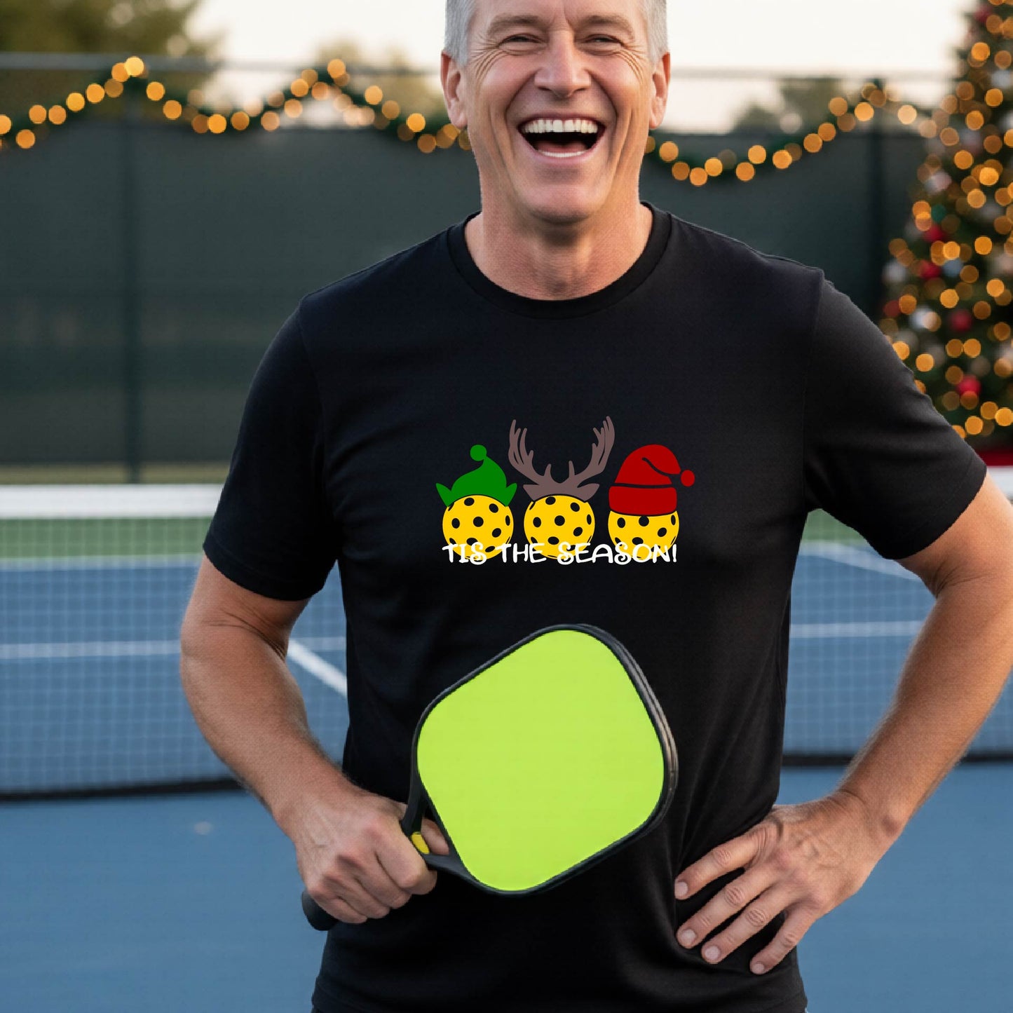 A man wearing a BLACK graphic short-sleeve t-shirt with a Christmas-themed design of three pickleballs wearing holiday hats and the text 'TIS THE SEASON!'. The background shows a festive blurred Christmas tree and a pickleball court.