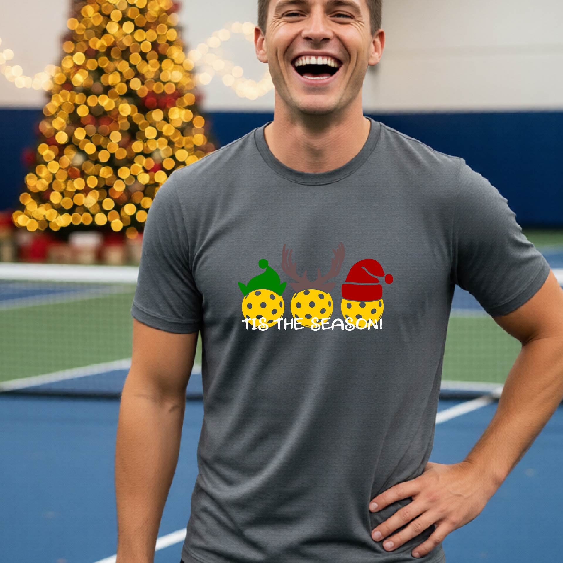 A man wearing a gray graphic short-sleeve t-shirt with a Christmas-themed design of three pickleballs wearing holiday hats and the text 'TIS THE SEASON!'. The background shows a festive blurred Christmas tree and a pickleball court.