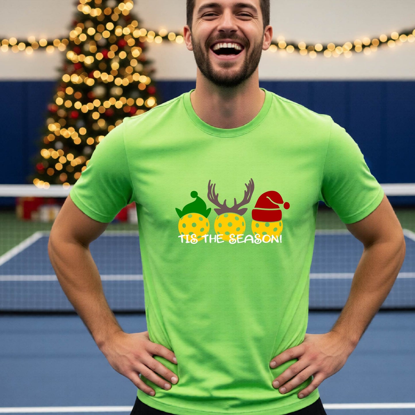 A man wearing a bright lime green graphic short-sleeve t-shirt with a Christmas-themed design of three pickleballs wearing holiday hats and the text 'TIS THE SEASON!'. The background shows a festive blurred Christmas tree and a pickleball court.