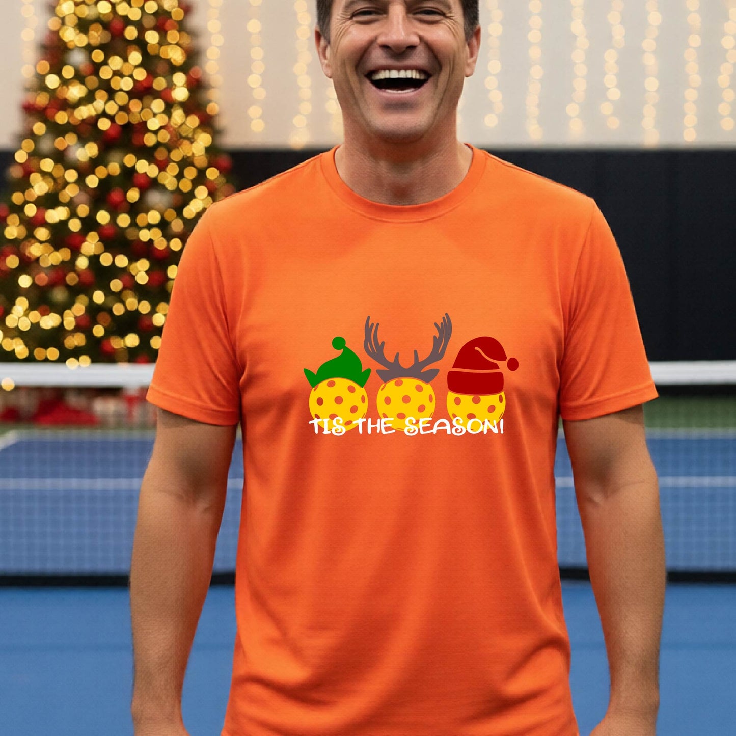 A man wearing a bright orange graphic short-sleeve t-shirt with a Christmas-themed design of three pickleballs wearing holiday hats and the text 'TIS THE SEASON!'. The background shows a festive blurred Christmas tree and a pickleball court.