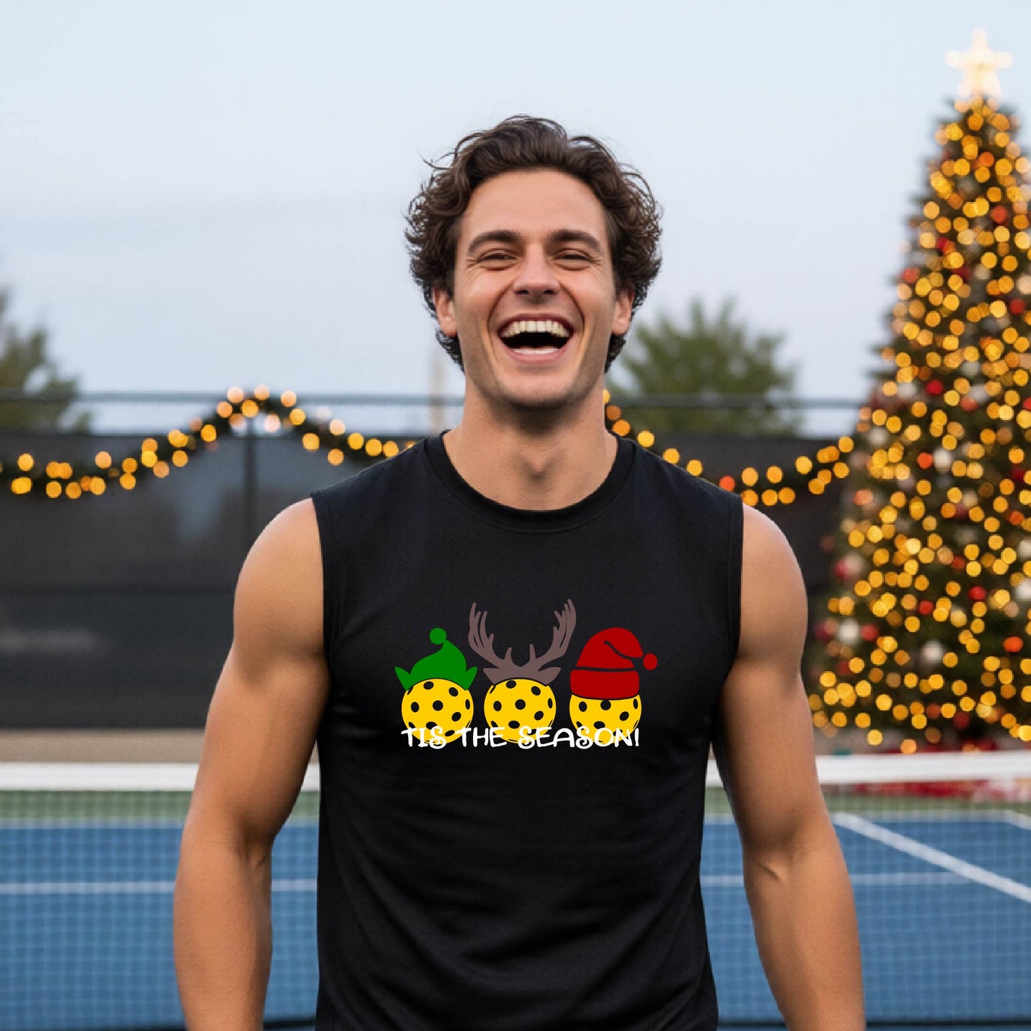 A smiling man wearing a black sleeveless athletic shirt with a pickleball Christmas-themed graphic of three pickleballs and the text 'TIS THE SEASON!' stands in front of a blurred background featuring a lit Christmas tree and a pickleball court.