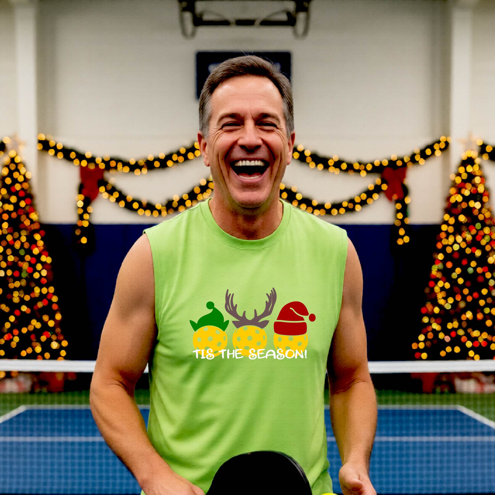 A smiling man wearing a lime green sleeveless athletic shirt with a pickleball Christmas-themed graphic of three pickleballs and the text 'TIS THE SEASON!' stands in front of a blurred background featuring a lit Christmas tree and a pickleball court.