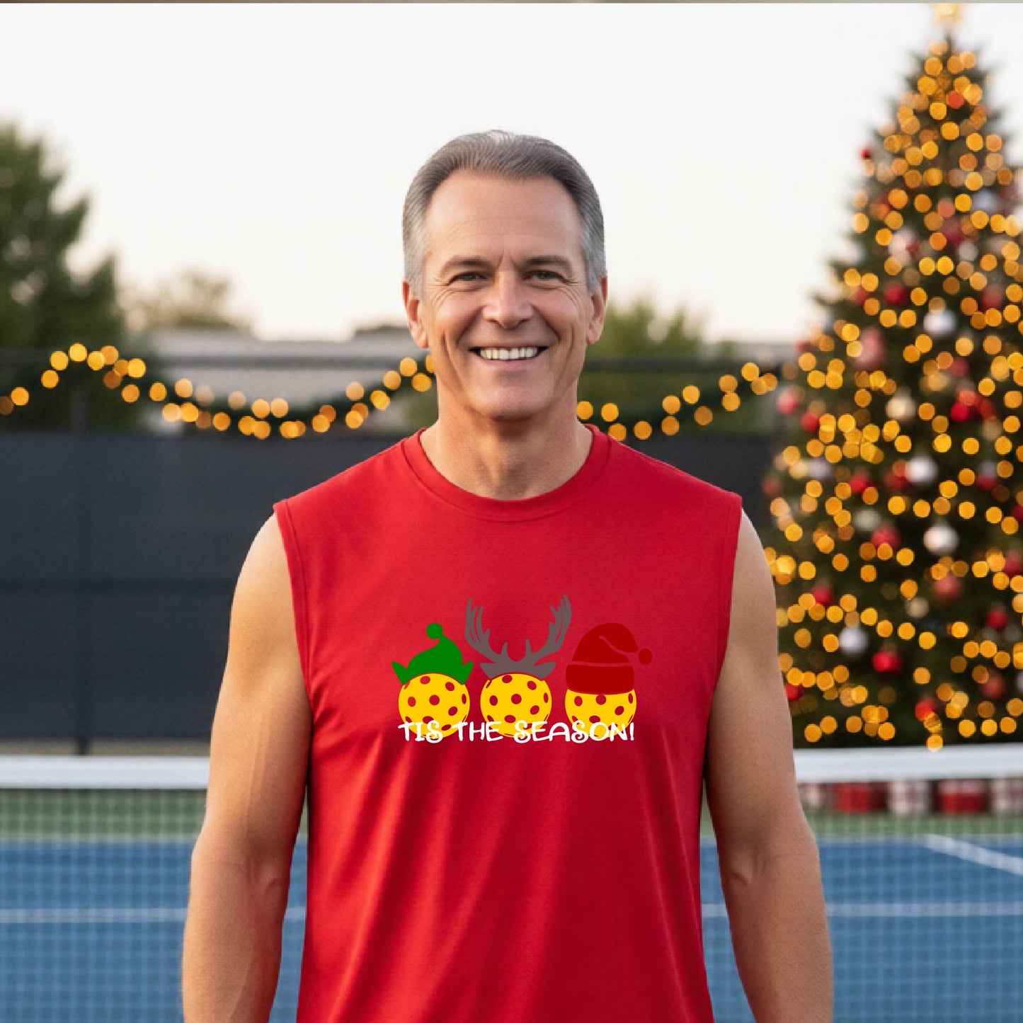 A smiling man wearing a red sleeveless athletic shirt with a pickleball Christmas-themed graphic of three pickleballs and the text 'TIS THE SEASON!' stands in front of a blurred background featuring a lit Christmas tree and a pickleball court.