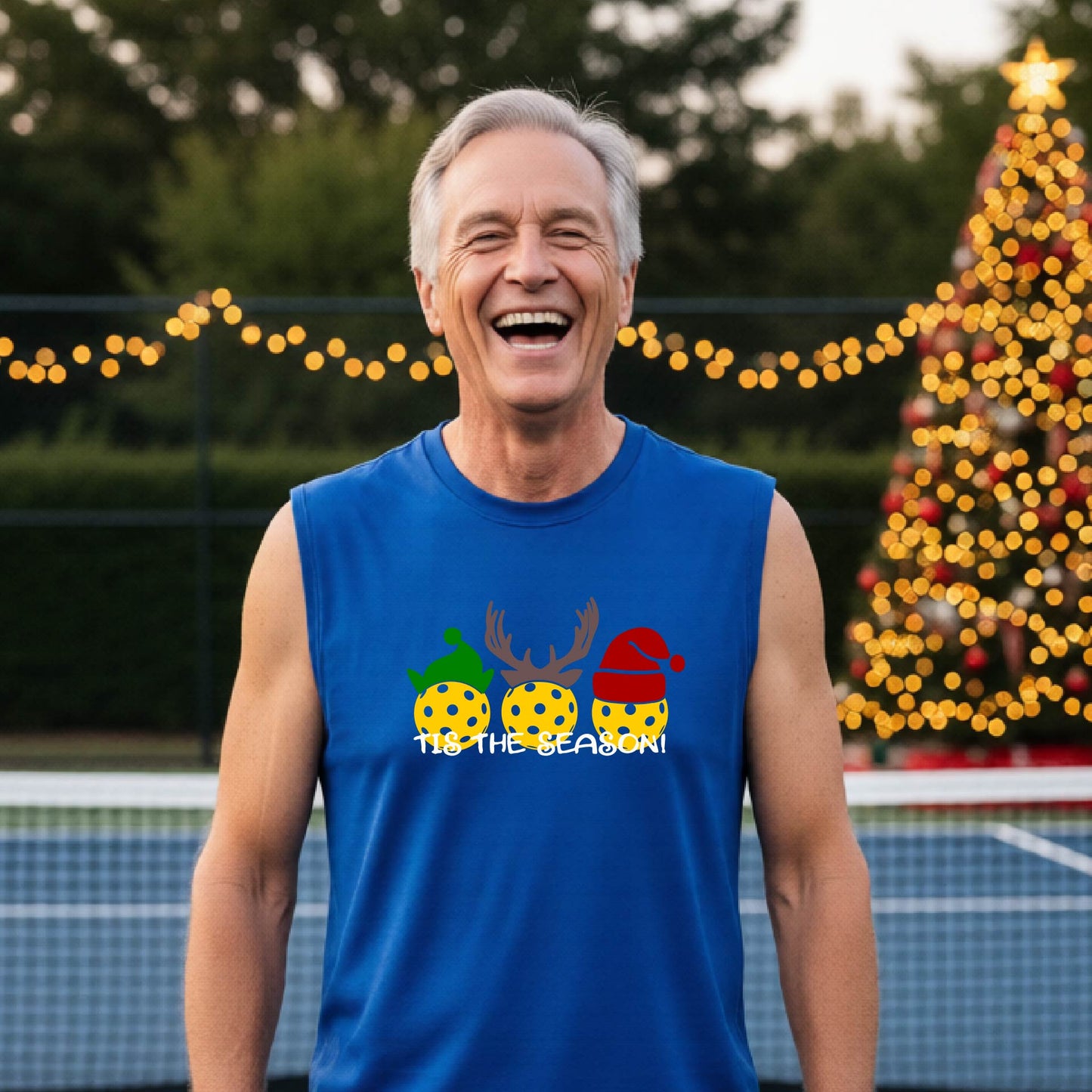 A smiling man wearing a royal blue sleeveless athletic shirt with a pickleball Christmas-themed graphic of three pickleballs and the text 'TIS THE SEASON!' stands in front of a blurred background featuring a lit Christmas tree and a pickleball court.