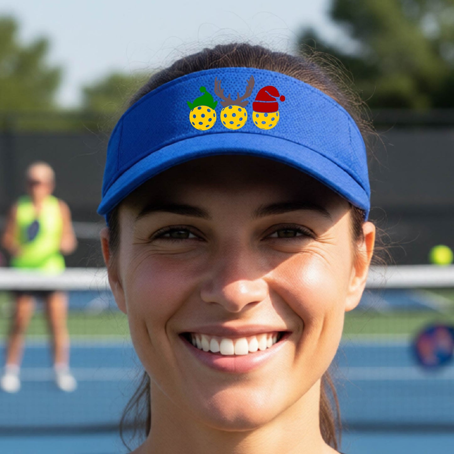 A royal blue visor with a pickleball Christmas graphic of three pickleballs wearing a santa hat, elf hat and antlers. 