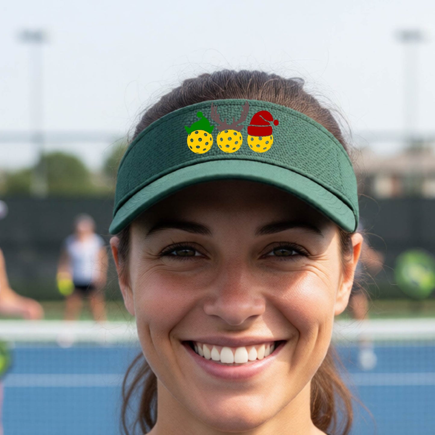 A green visor with a pickleball Christmas graphic of three pickleballs wearing a santa hat, elf hat and antlers. 