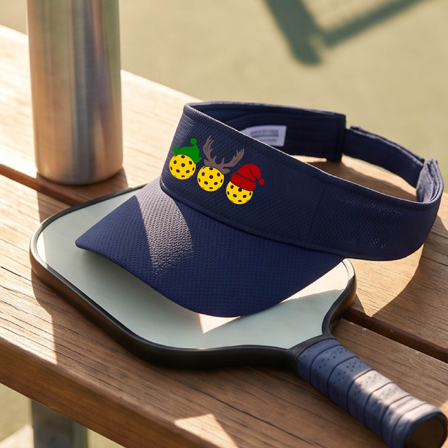 A navy blue visor with a pickleball Christmas graphic of three pickleballs wearing a santa hat, elf hat and antlers. 