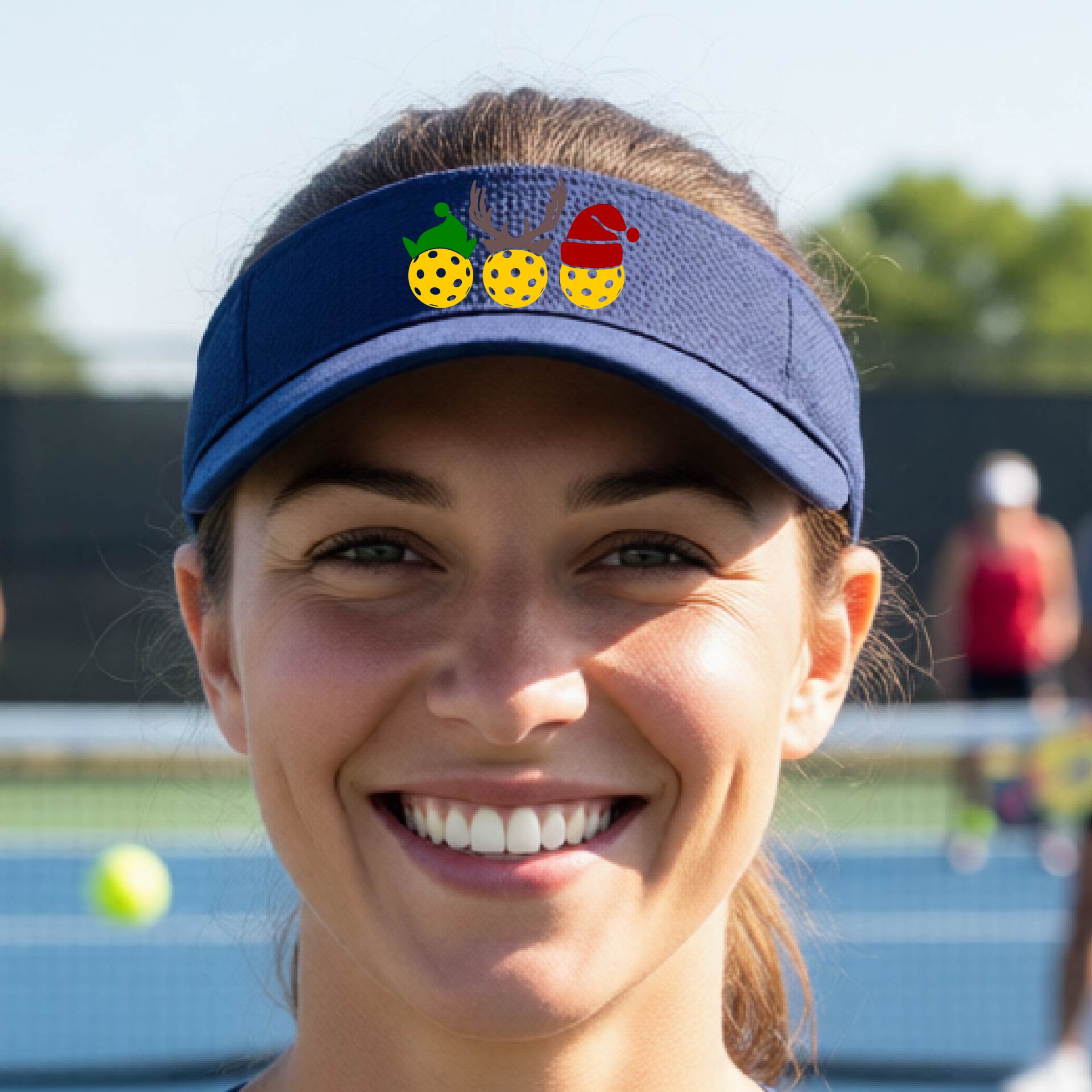 A navy blue visor with a pickleball Christmas graphic of three pickleballs wearing a santa hat, elf hat and antlers. 