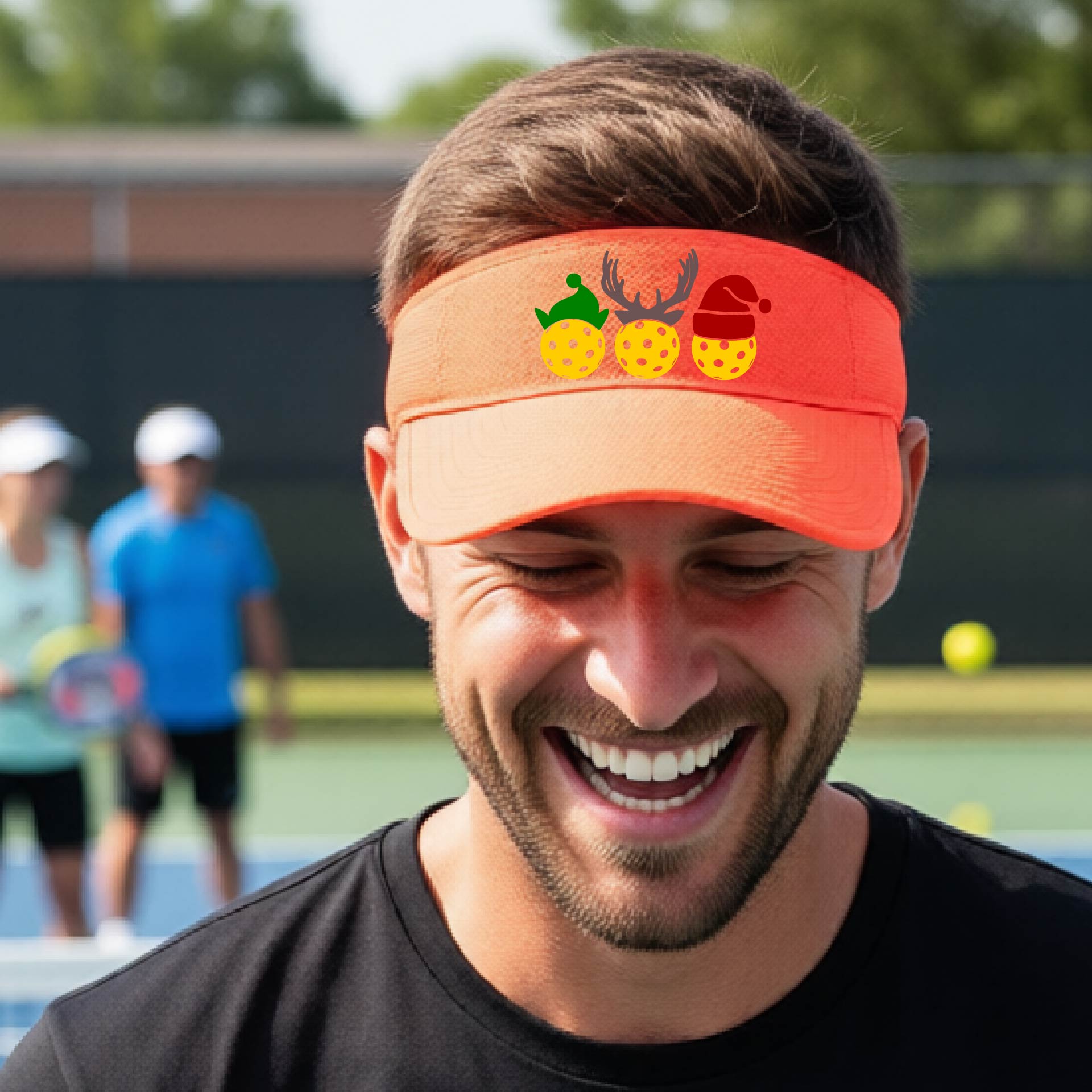 An orange visor with a pickleball Christmas graphic of three pickleballs wearing a santa hat, elf hat and antlers. 