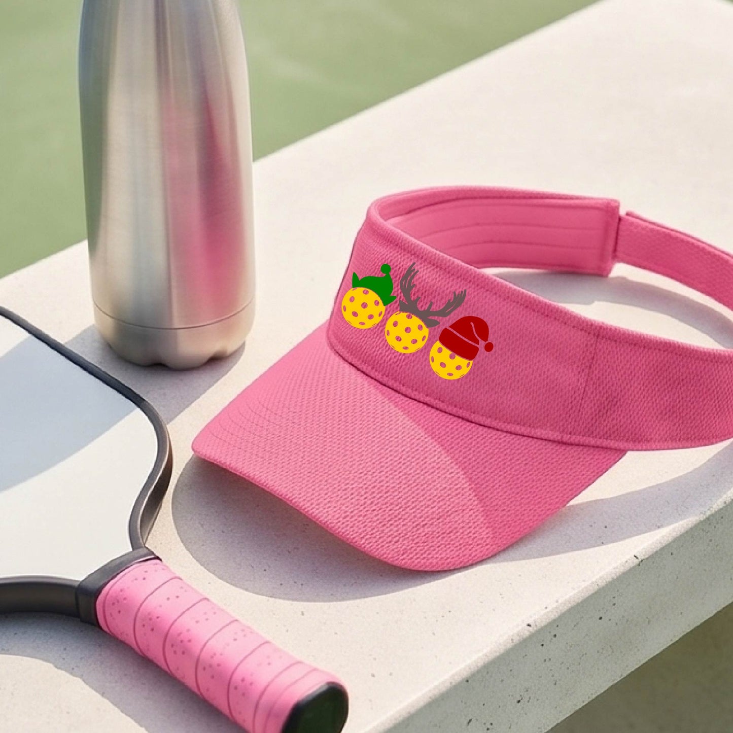 A pink visor with a pickleball Christmas graphic of three pickleballs wearing a santa hat, elf hat and antlers. 