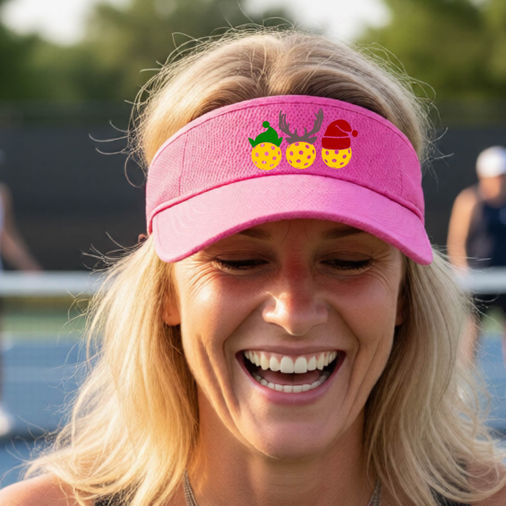 A pink visor with a pickleball Christmas graphic of three pickleballs wearing a santa hat, elf hat and antlers. 