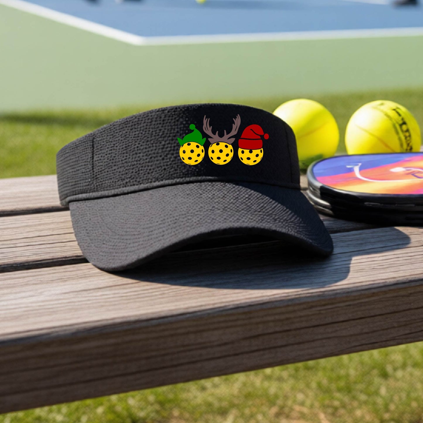 A black visor with a pickleball Christmas graphic of three pickleballs wearing a santa hat, elf hat and antlers. 