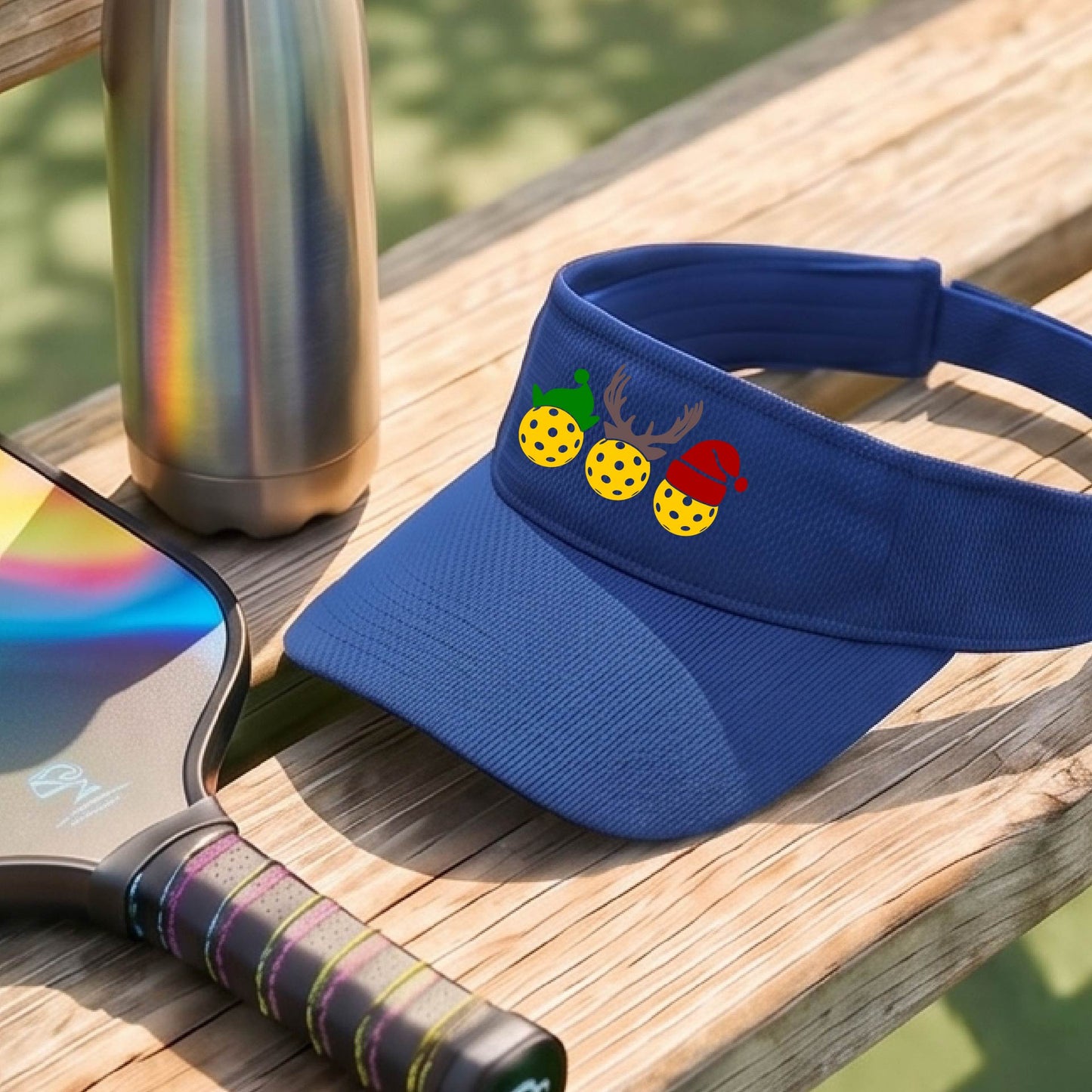 A royal blue visor with a pickleball Christmas graphic of three pickleballs wearing a santa hat, elf hat and antlers. 