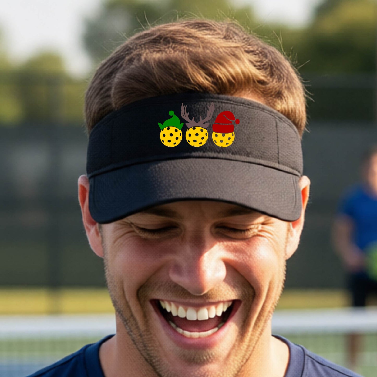 A black visor with a pickleball Christmas graphic of three pickleballs wearing a santa hat, elf hat and antlers. 