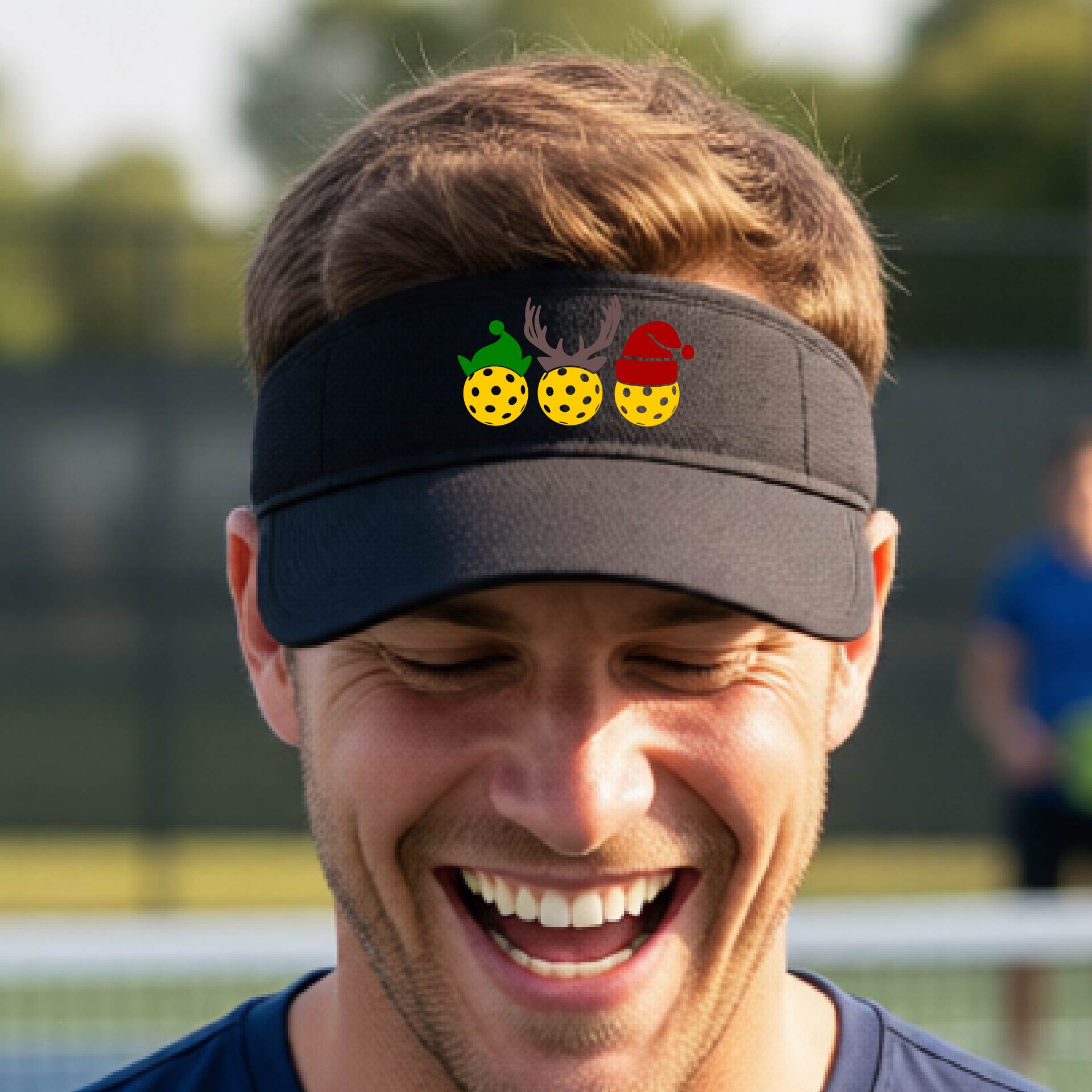 A black visor with a pickleball Christmas graphic of three pickleballs wearing a santa hat, elf hat and antlers. 