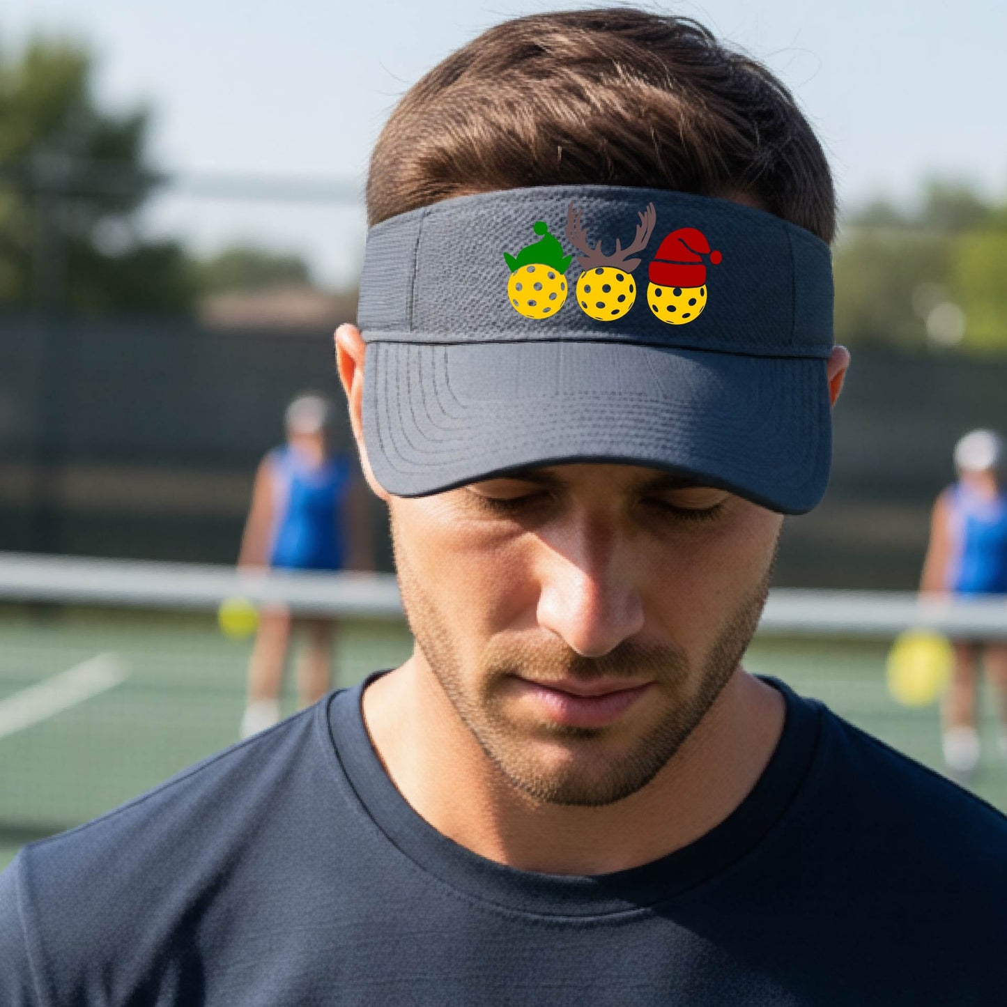 A graphite gray visor with a pickleball Christmas graphic of three pickleballs wearing a santa hat, elf hat and antlers. 