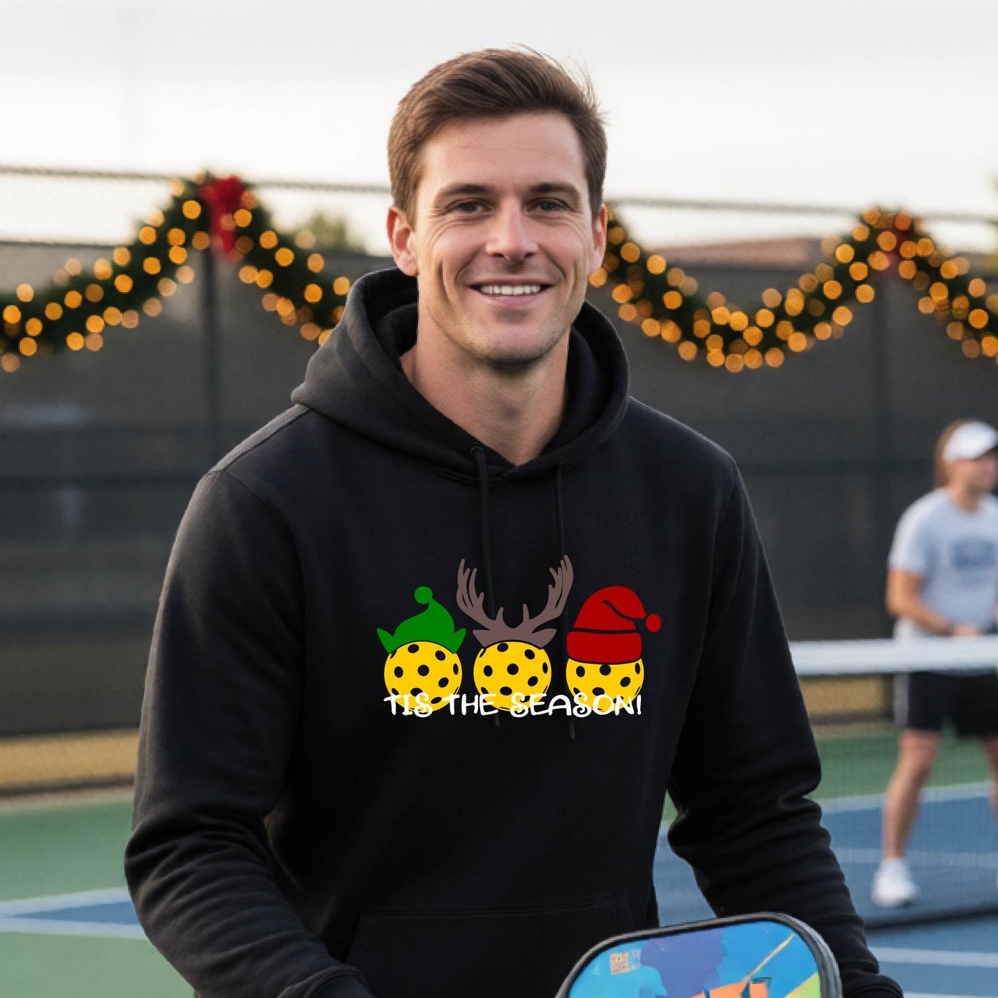 A man wearing a black hoodie with a Christmas-themed graphic of three pickleballs characters wearing green, brown, and red hats, with the text 'TIS THE SEASON!' underneath.