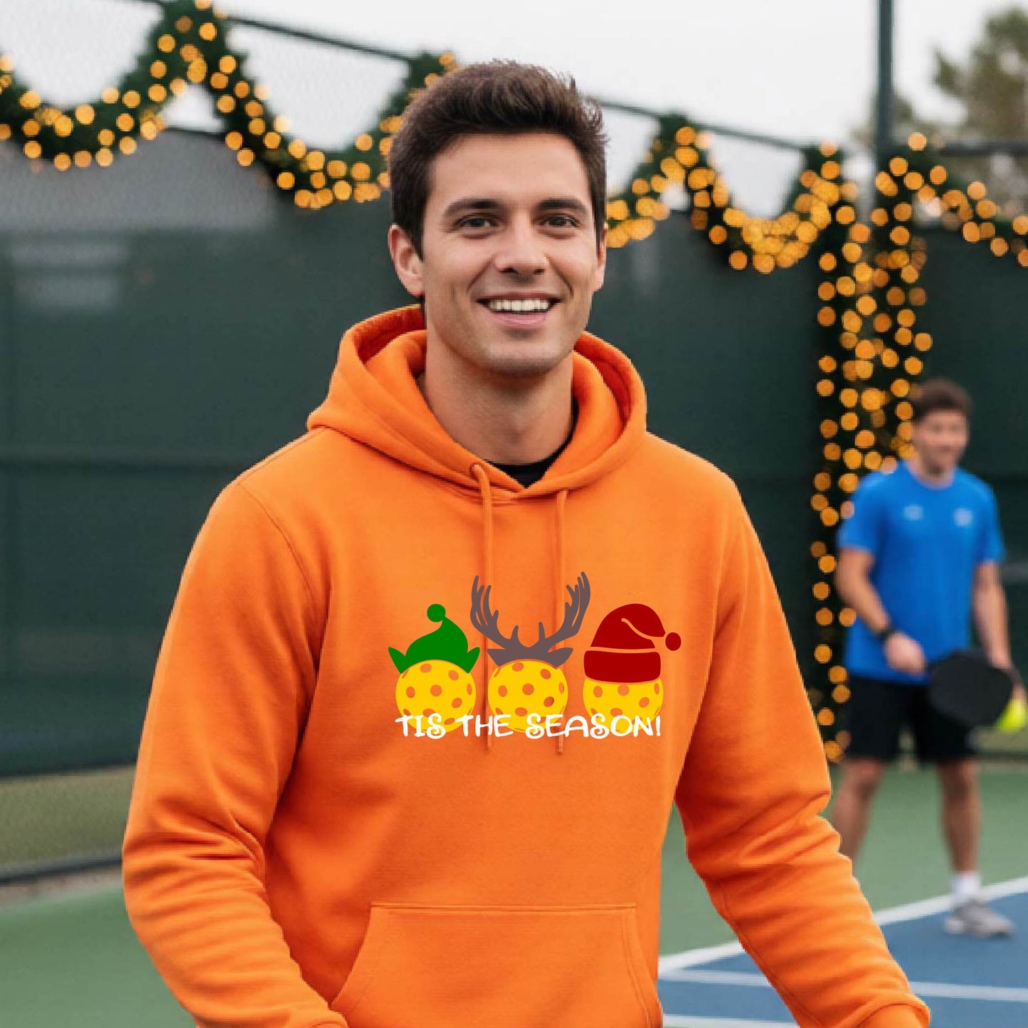A man wearing an orange hoodie with a Christmas-themed graphic of three pickleballs characters wearing green, brown, and red hats, with the text 'TIS THE SEASON!' underneath.