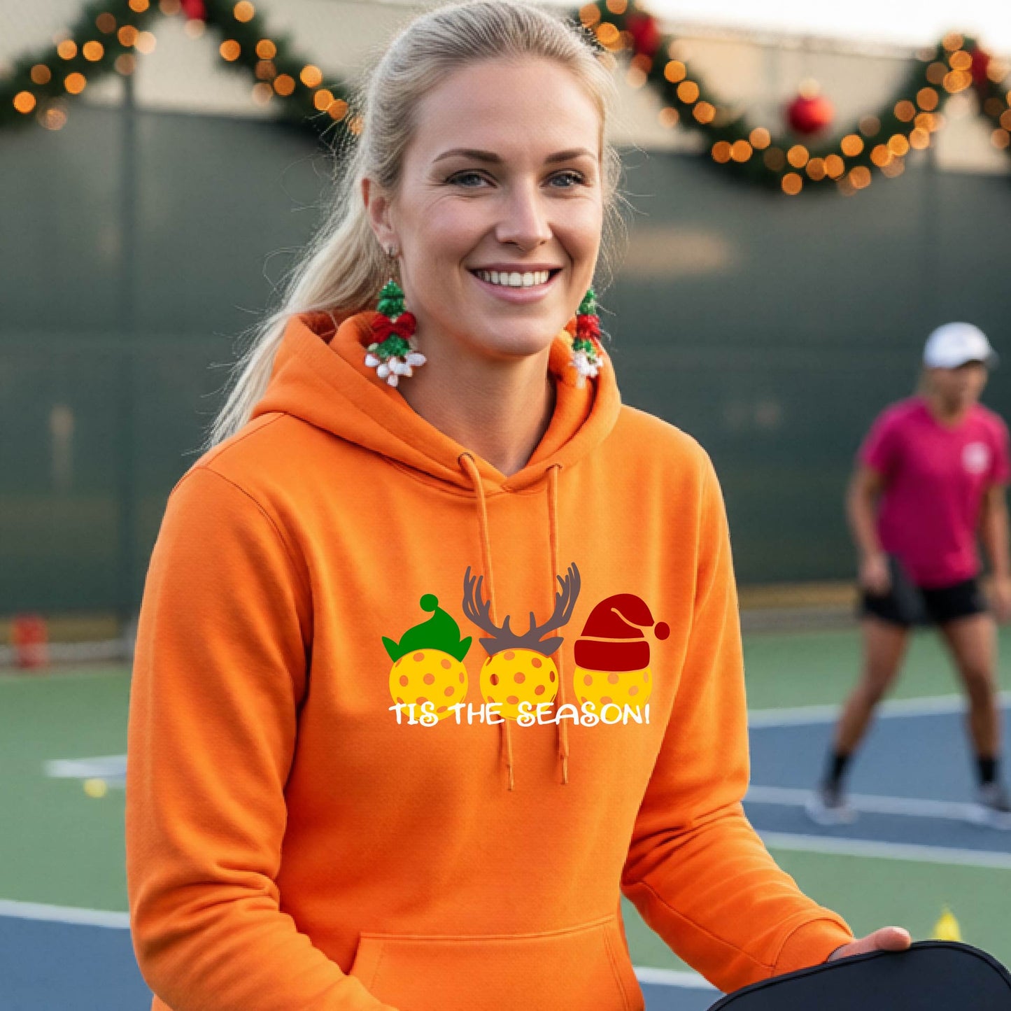 A woman wearing an orange hoodie with a Christmas-themed graphic of three pickleballs characters wearing green, brown, and red hats, with the text 'TIS THE SEASON!' underneath.