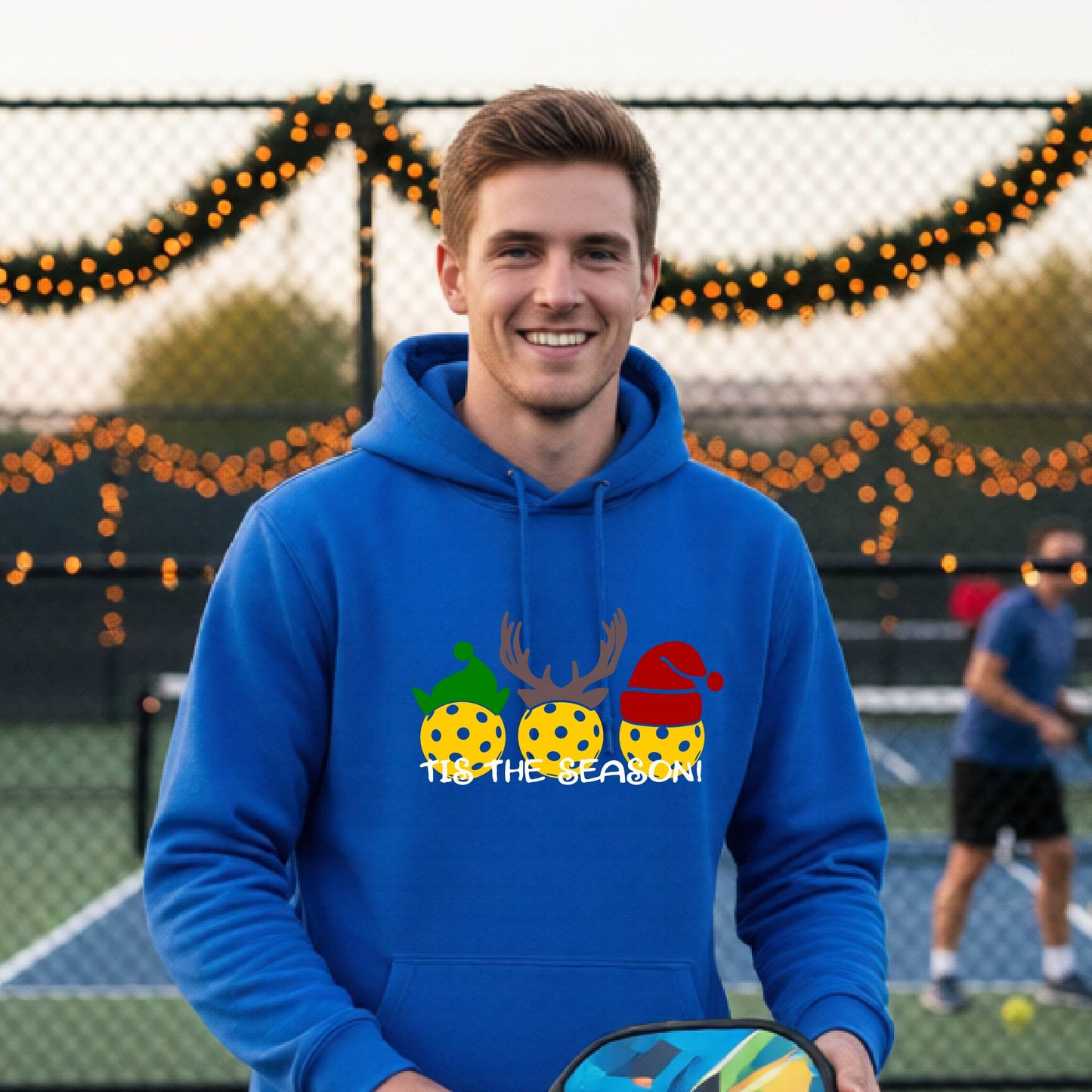 A man wearing a royal blue hoodie with a Christmas-themed graphic of three pickleballs characters wearing green, brown, and red hats, with the text 'TIS THE SEASON!' underneath.
