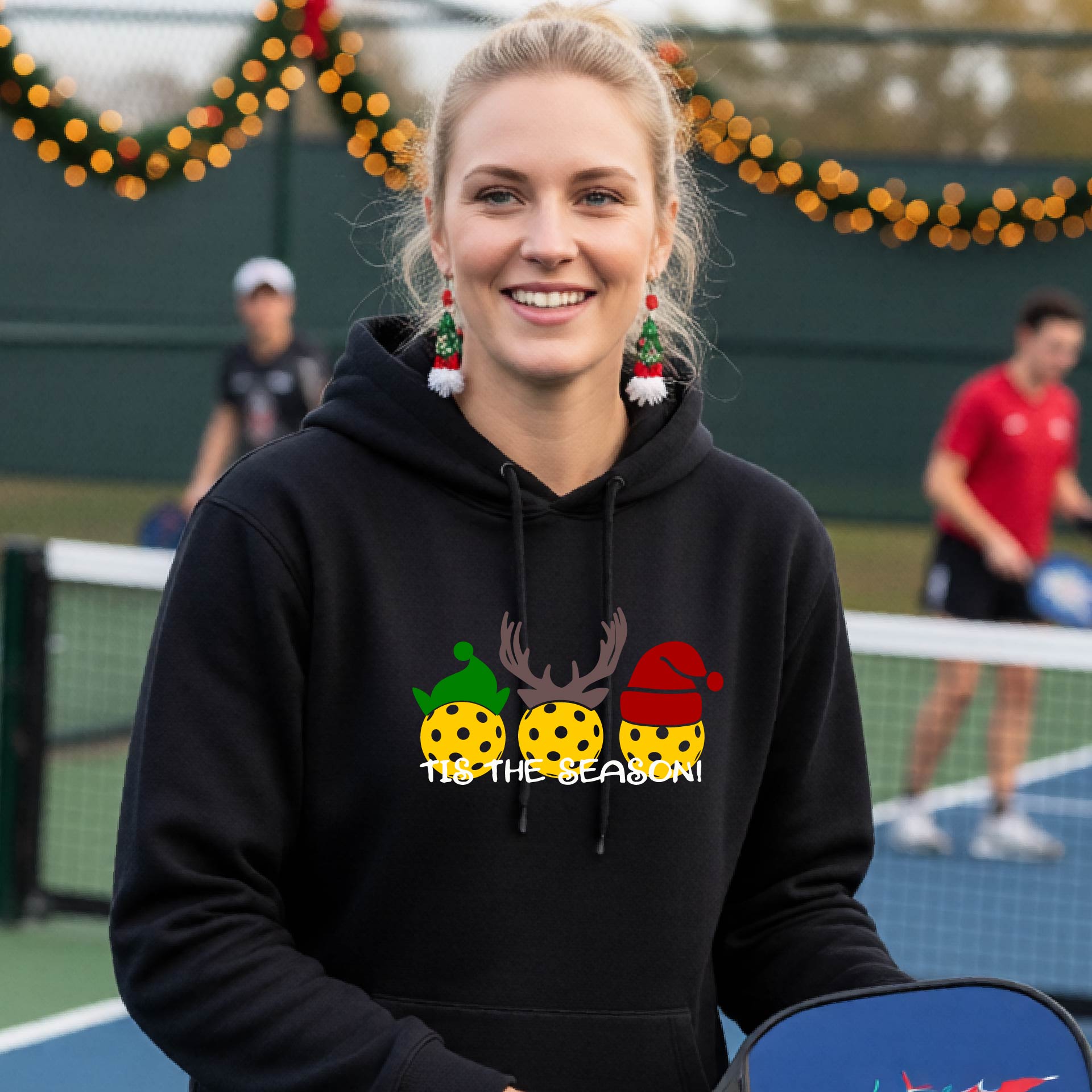 A woman wearing a black hoodie with a Christmas-themed graphic of three pickleballs characters wearing green, brown, and red hats, with the text 'TIS THE SEASON!' underneath.