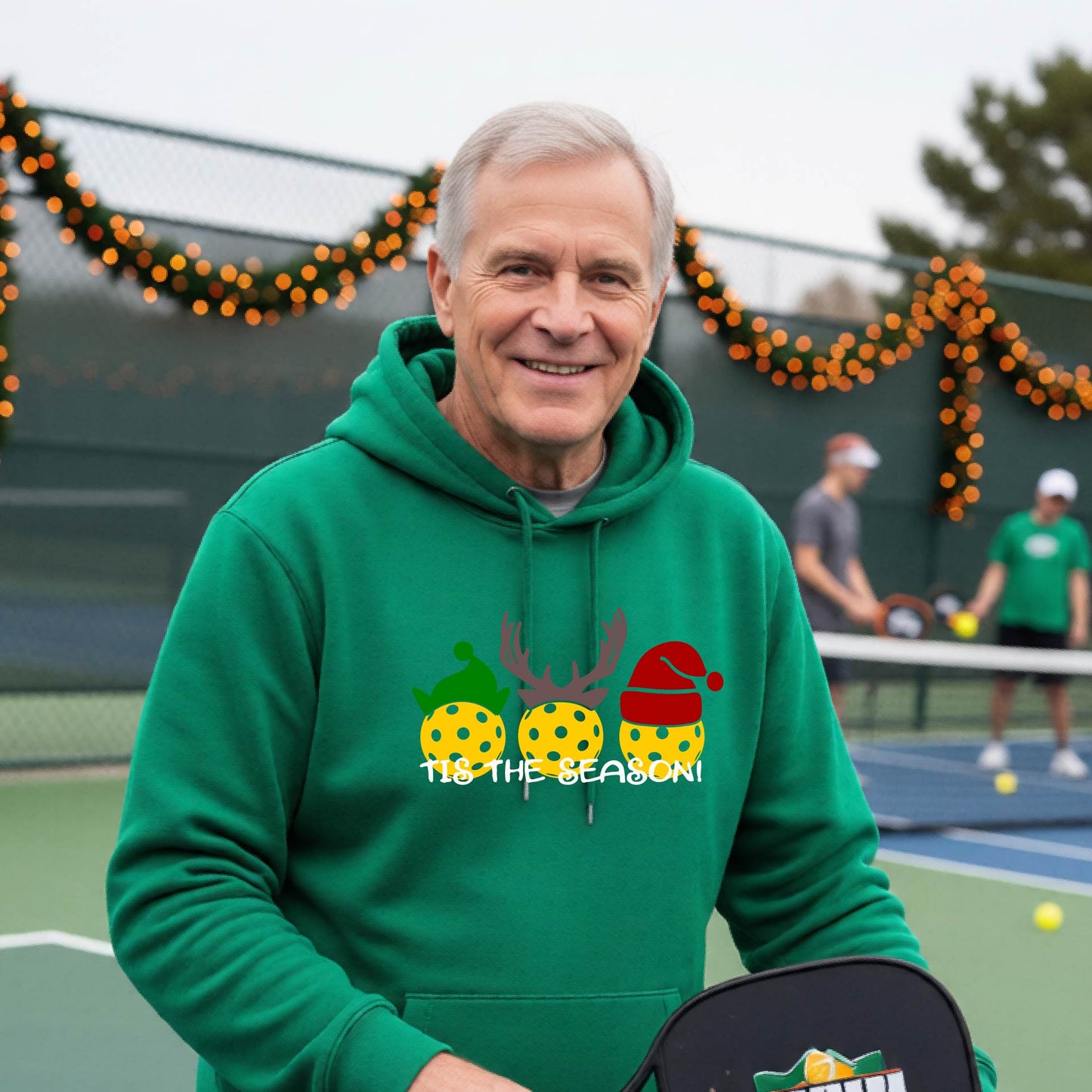 A man wearing a green hoodie with a Christmas-themed graphic of three pickleballs characters wearing green, brown, and red hats, with the text 'TIS THE SEASON!' underneath.