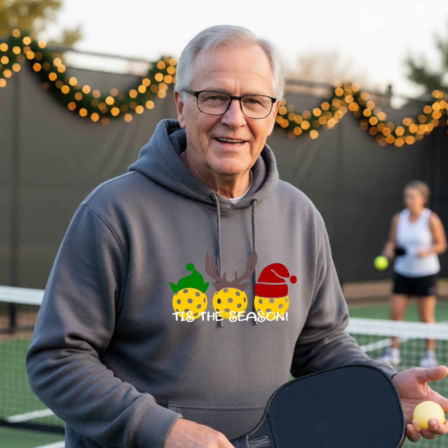 A man wearing a gray hoodie with a Christmas-themed graphic of three pickleballs characters wearing green, brown, and red hats, with the text 'TIS THE SEASON!' underneath.