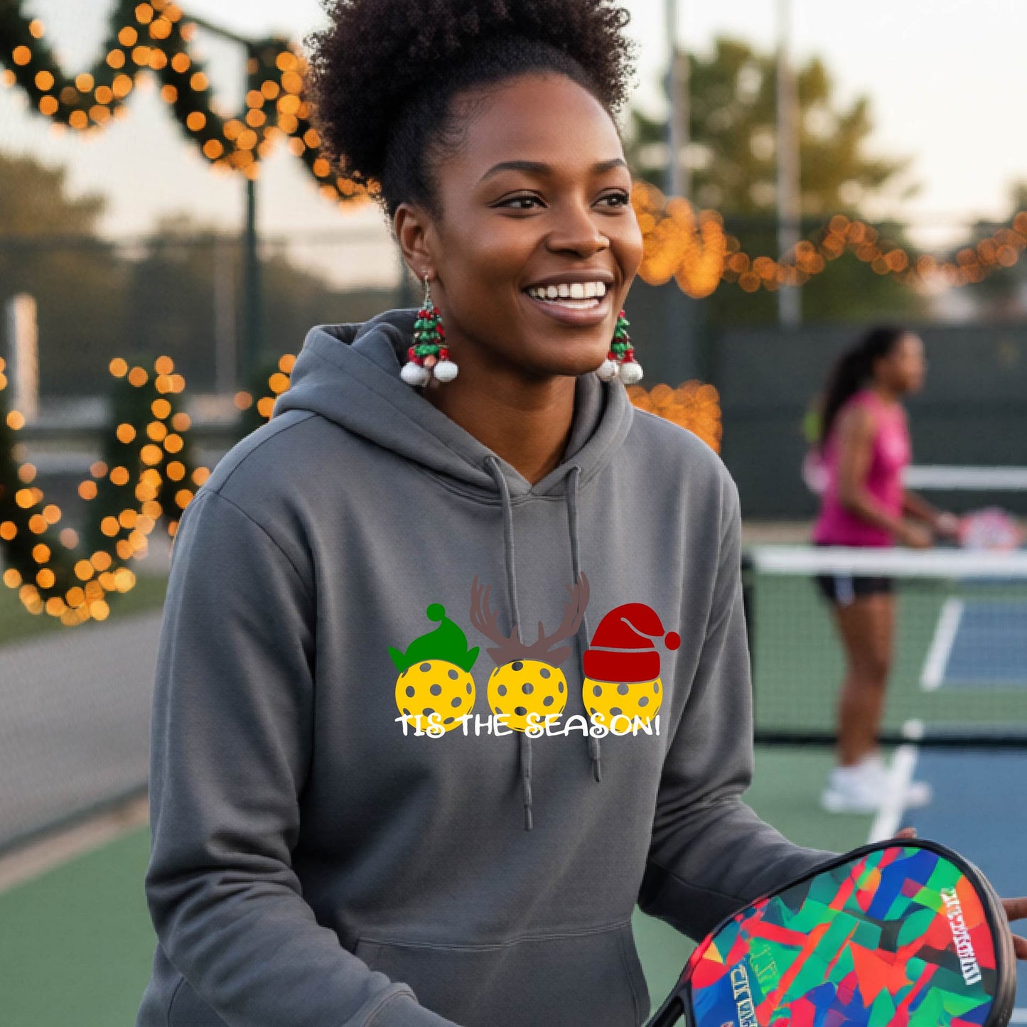 A woman wearing a black hoodie with a Christmas-themed graphic of three pickleballs characters wearing green, brown, and red hats, with the text 'TIS THE SEASON!' underneath.