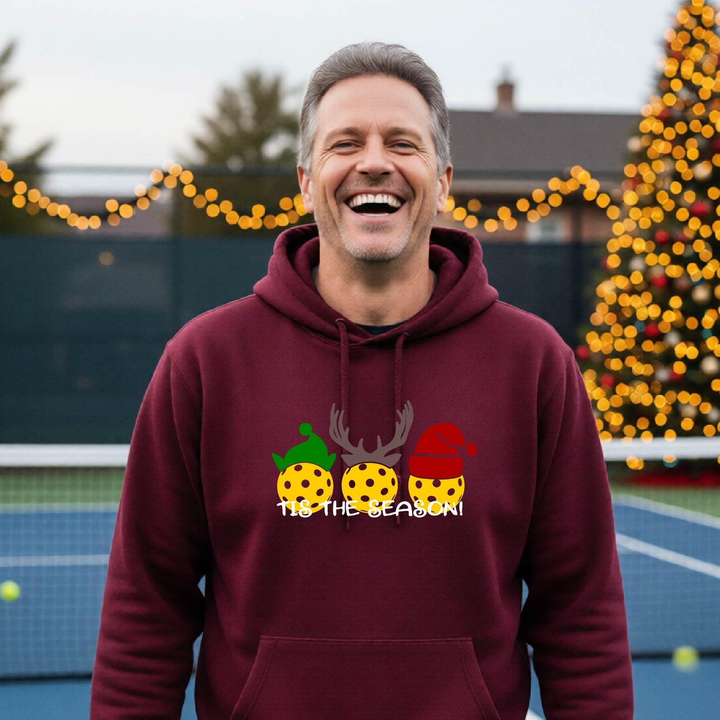 A man wearing a maroon hoodie with a Christmas-themed graphic of three pickleballs characters wearing green, brown, and red hats, with the text 'TIS THE SEASON!' underneath.