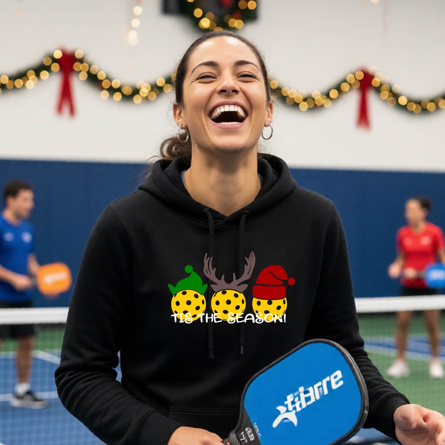 A woman laughing on an indoor pickleball court wearing a black hoodie with a festive Christmas-pickleball themed graphic of two pickleball players in holiday outfits and the text 'TIS THE SEASON!'. 