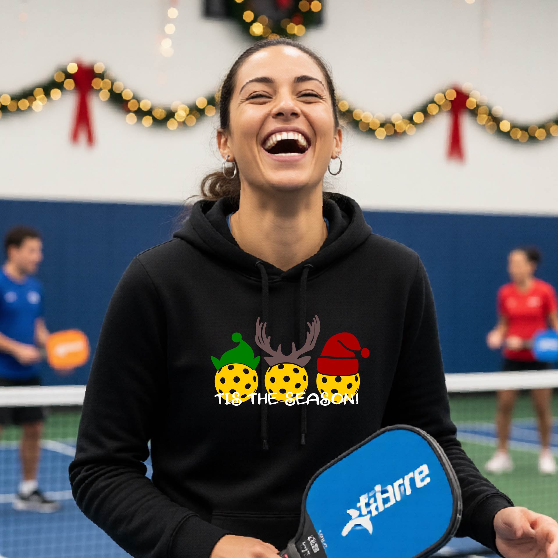A woman laughing on an indoor pickleball court wearing a black hoodie with a festive Christmas-pickleball themed graphic of two pickleball players in holiday outfits and the text 'TIS THE SEASON!'. 