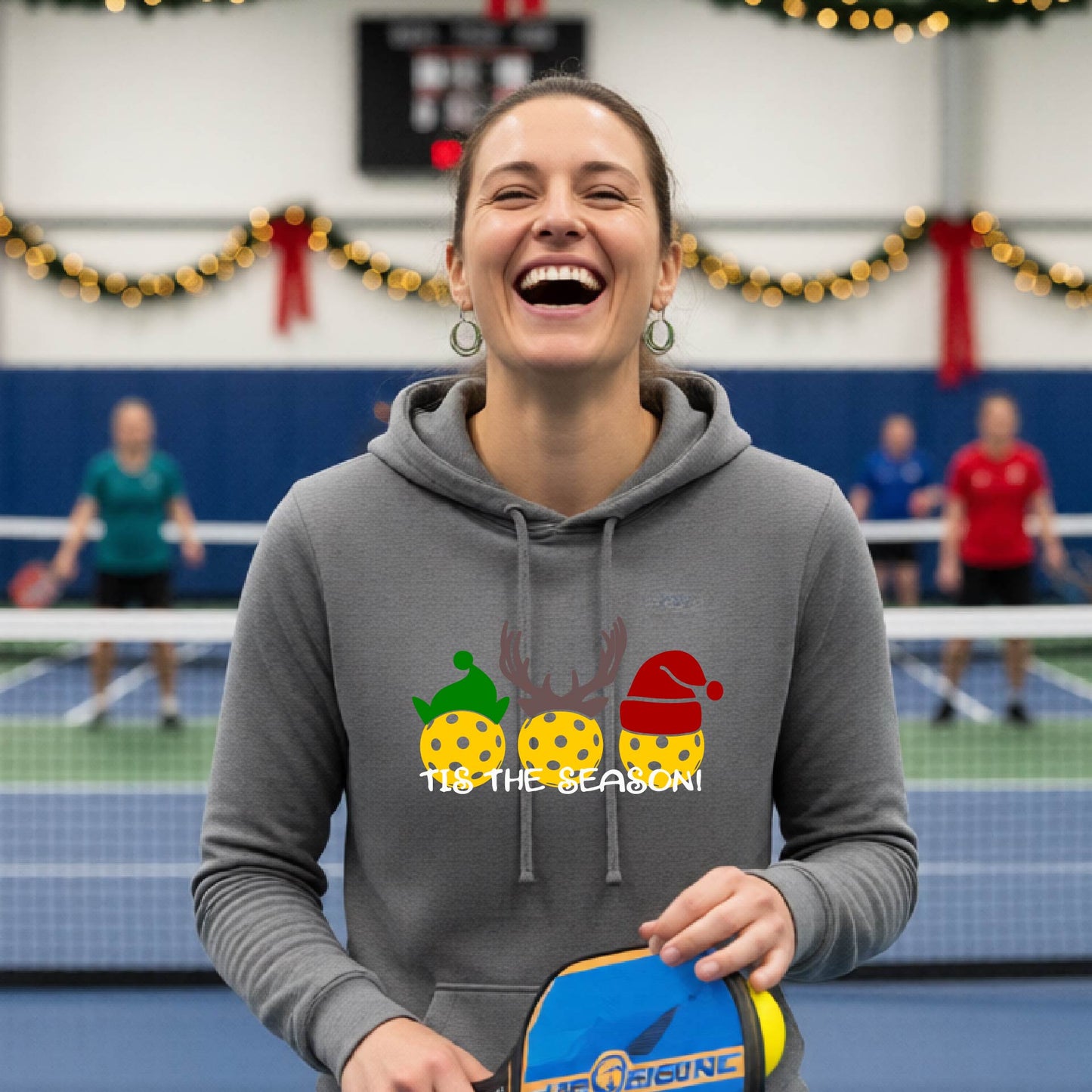 A woman laughing on an indoor pickleball court wearing a heather gray hoodie with a festive Christmas-pickleball themed graphic of two pickleball players in holiday outfits and the text 'TIS THE SEASON!'. 