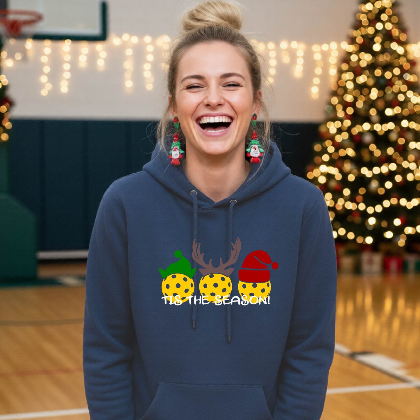 A woman laughing on an indoor pickleball court wearing a heather navy hoodie with a festive Christmas-pickleball themed graphic of two pickleball players in holiday outfits and the text 'TIS THE SEASON!'. 
