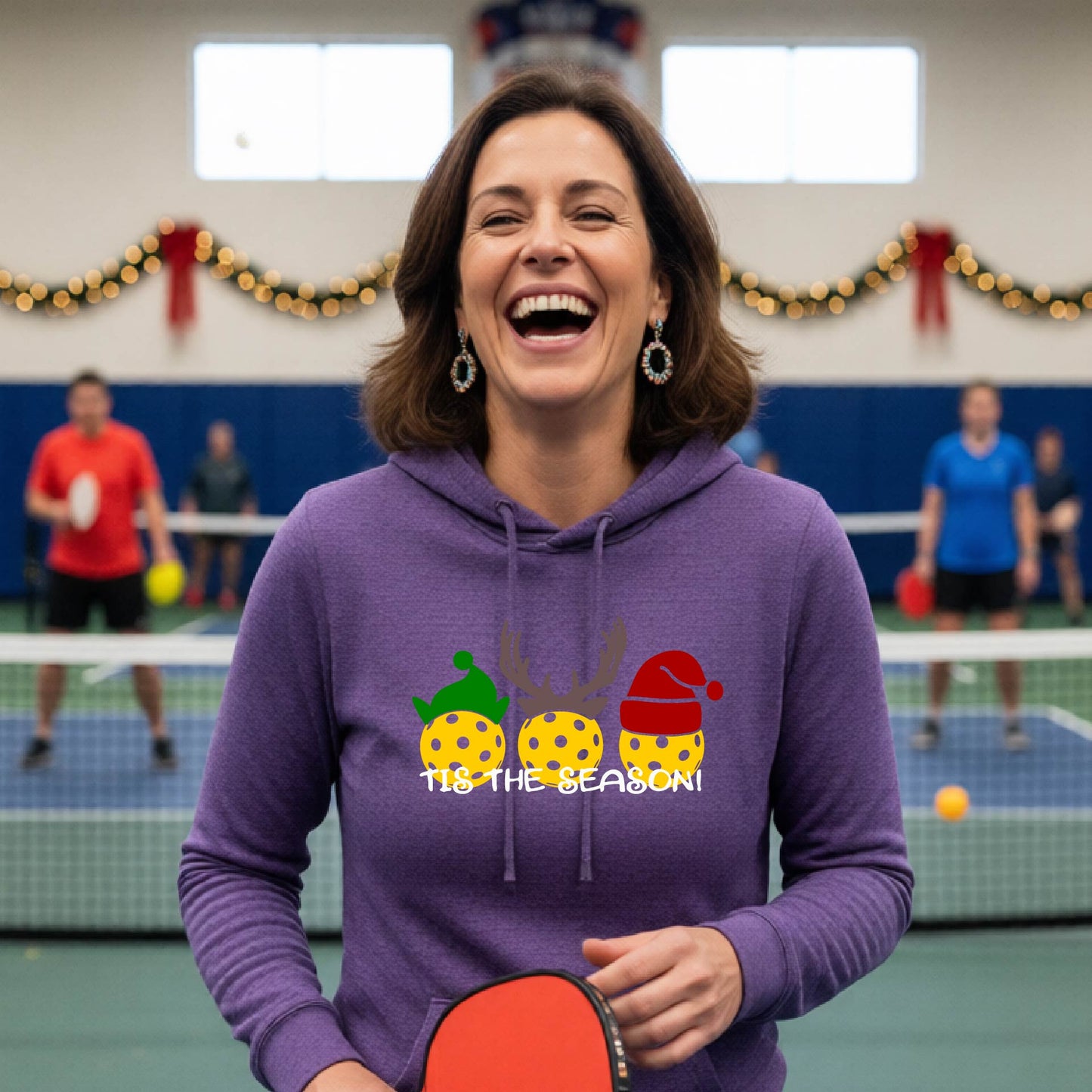 A woman laughing on an indoor pickleball court wearing a heather purple hoodie with a festive Christmas-pickleball themed graphic of two pickleball players in holiday outfits and the text 'TIS THE SEASON!'. 
