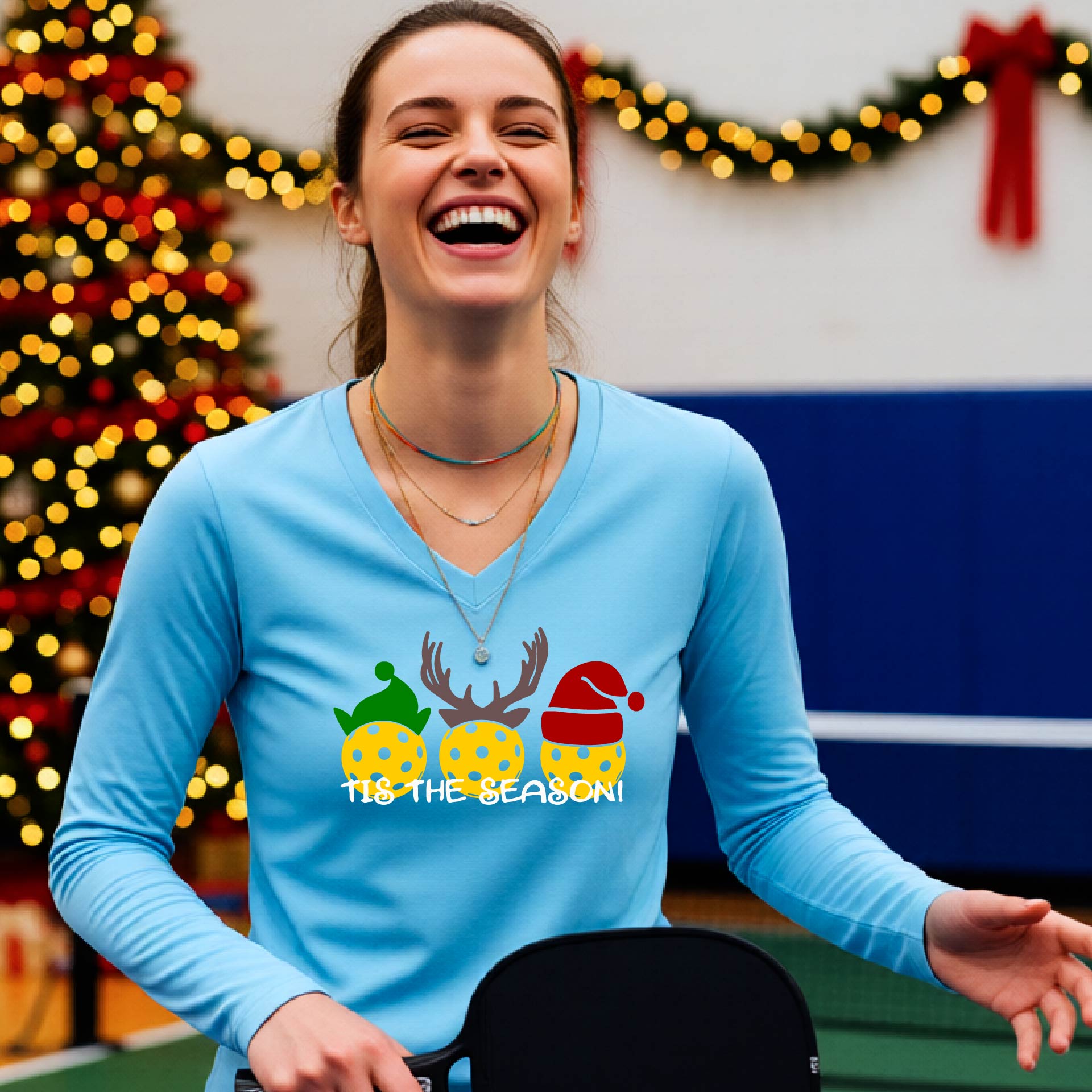 A woman smiling in a gym wearing a light blue long-sleeve V-neck shirt with a festive graphic of three Pickleballs decorated like animals and the text 'TIS THE SEASON!'. A decorated Christmas tree and holiday lights are visible in the background.
