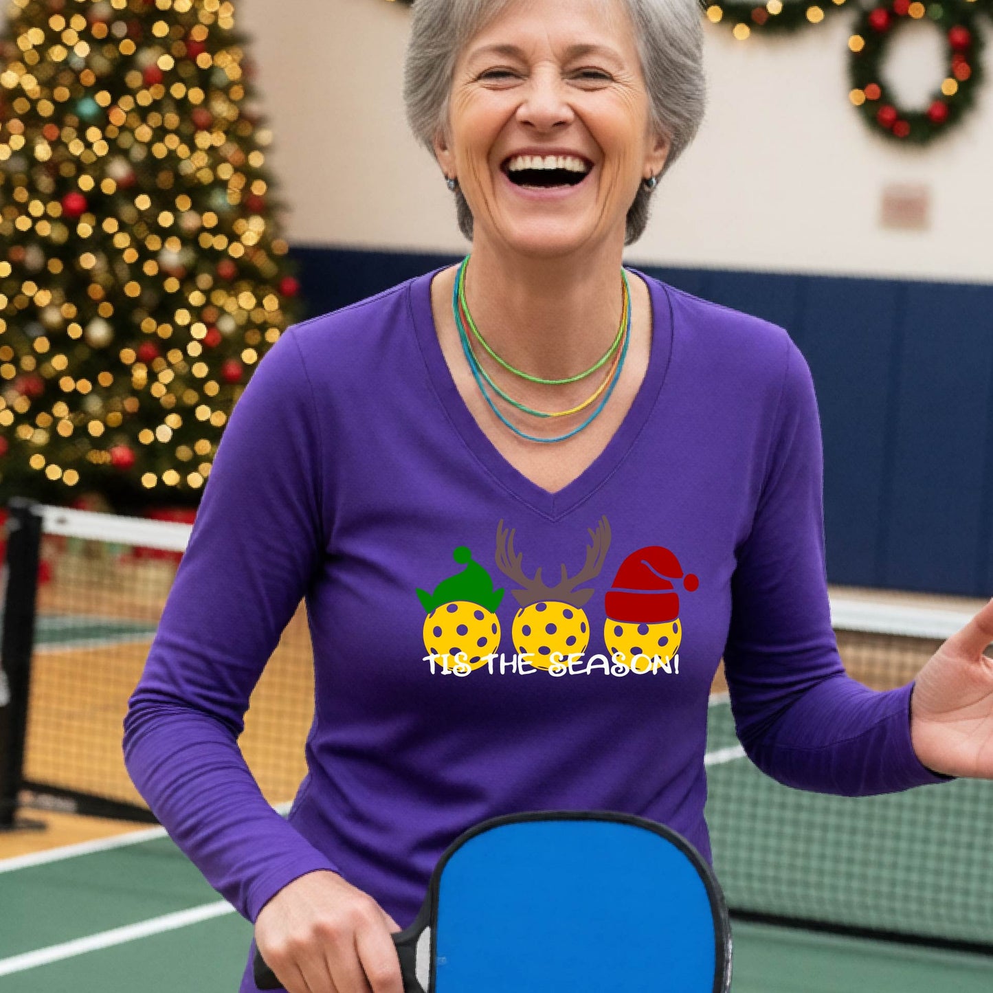 A woman smiling in a gym wearing a purple long-sleeve V-neck shirt with a festive graphic of three Pickleballs decorated like animals and the text 'TIS THE SEASON!'. A decorated Christmas tree and holiday lights are visible in the background.