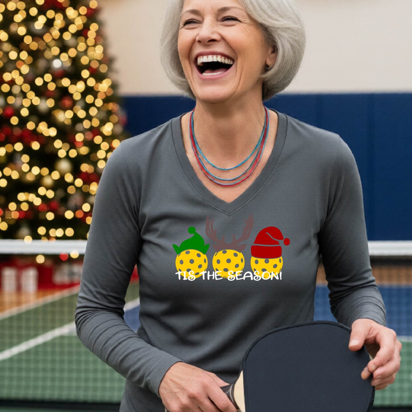 A woman smiling in a gym wearing a gray long-sleeve V-neck shirt with a festive graphic of three Pickleballs decorated like animals and the text 'TIS THE SEASON!'. A decorated Christmas tree and holiday lights are visible in the background.