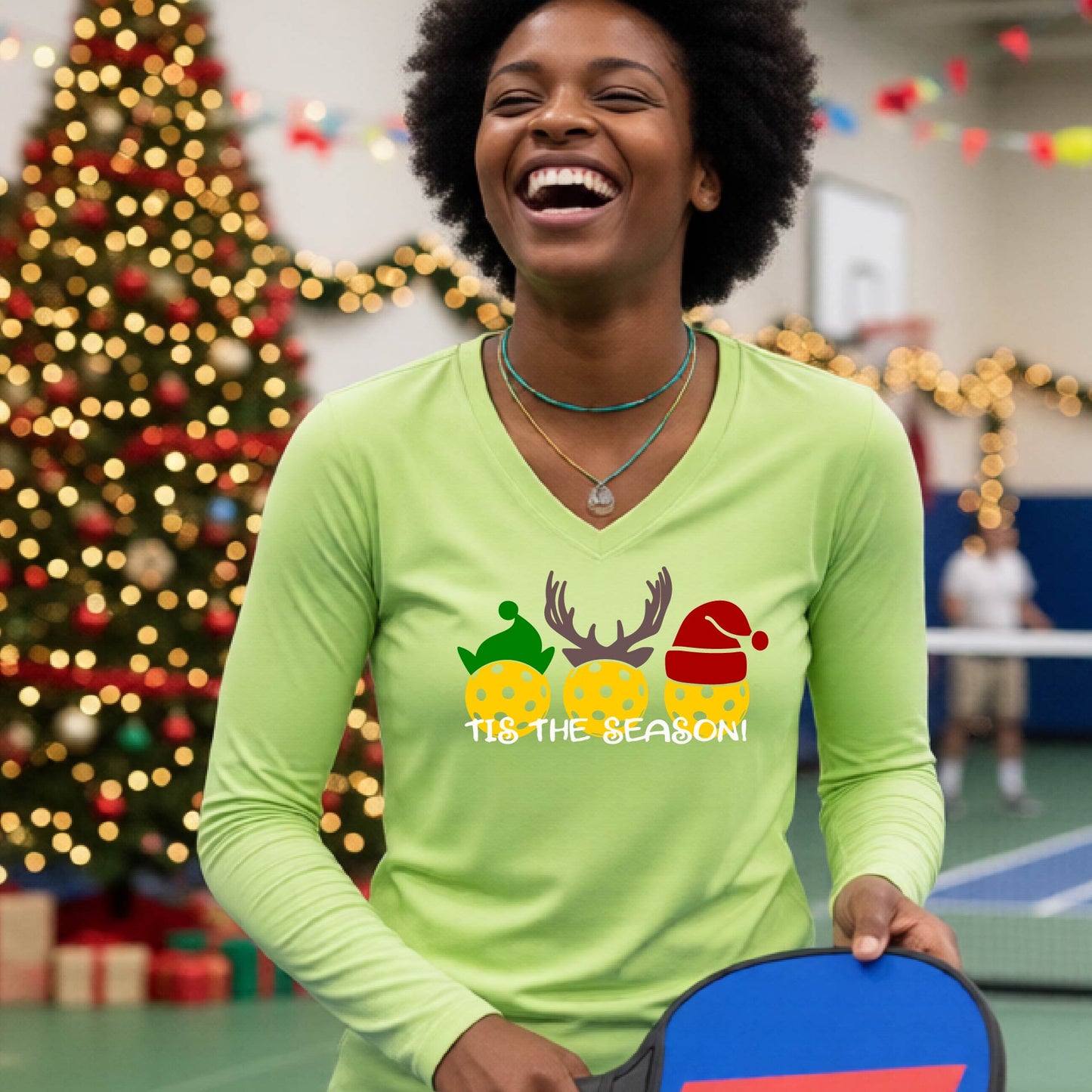 A woman smiling in a gym wearing a light green long-sleeve V-neck shirt with a festive graphic of three Pickleballs decorated like animals and the text 'TIS THE SEASON!'. A decorated Christmas tree and holiday lights are visible in the background.