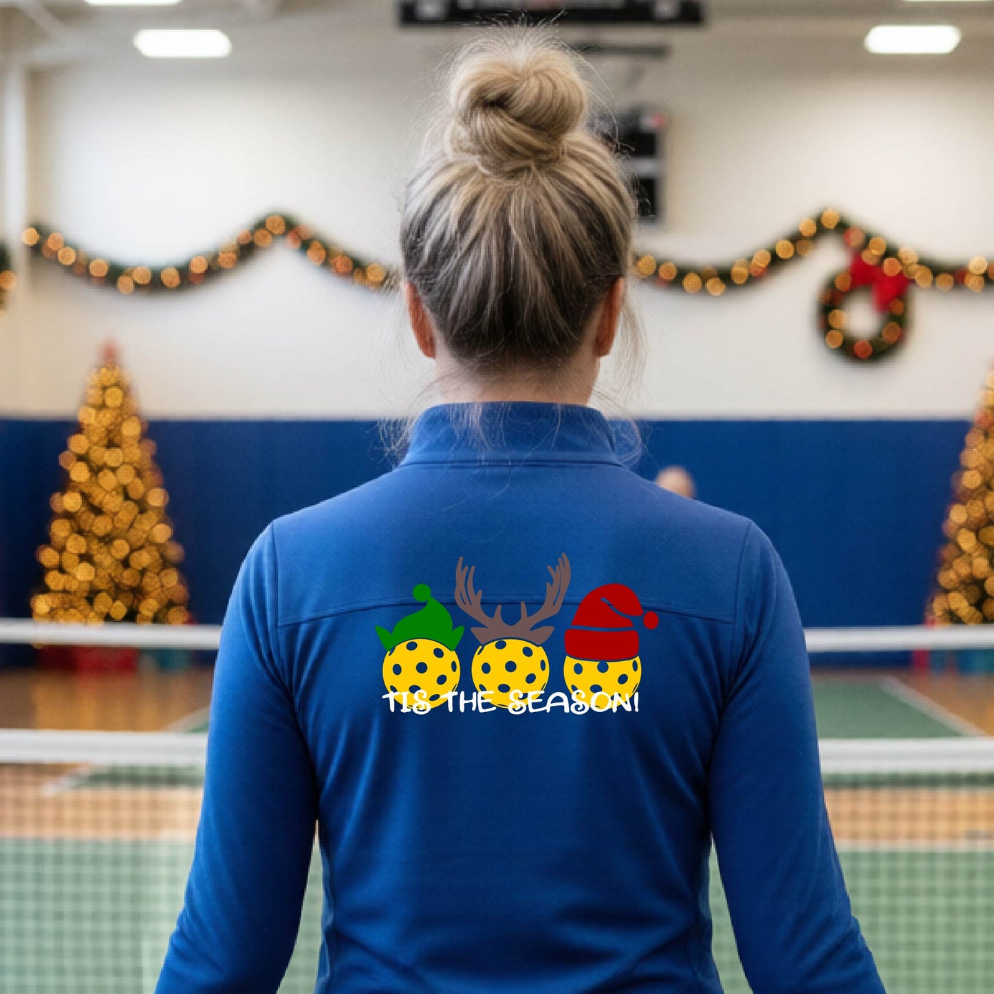 A woman viewed from behind wearing a royal blue long-sleeve athletic top with a Christmas-themed graphic on the back. The graphic shows three yellow picKlebalLS with green, brown, and red holiday patterns, along with white text reading "TIS THE SEASON"