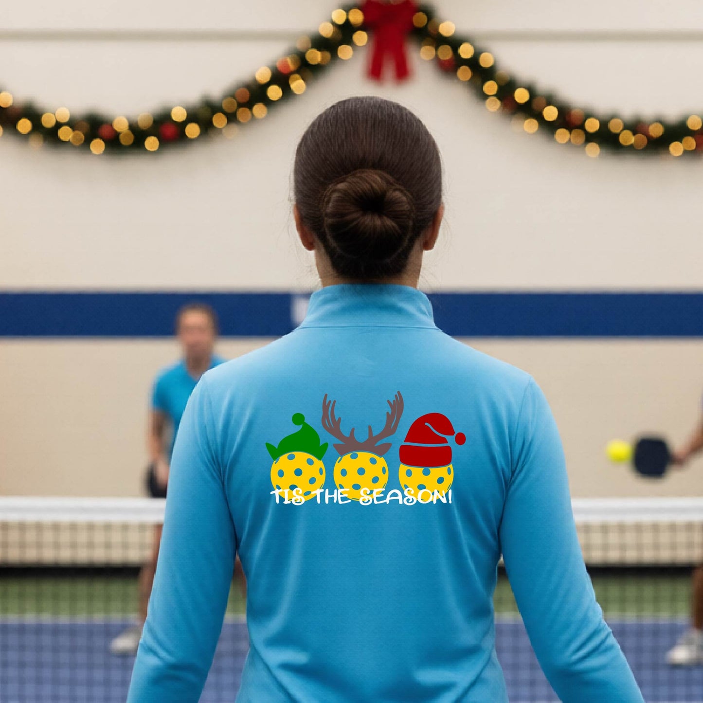 A woman viewed from behind wearing a light blue long-sleeve athletic top with a Christmas-themed graphic on the back. The graphic shows three yellow picKlebalLS with green, brown, and red holiday patterns, along with white text reading "TIS THE SEASON"