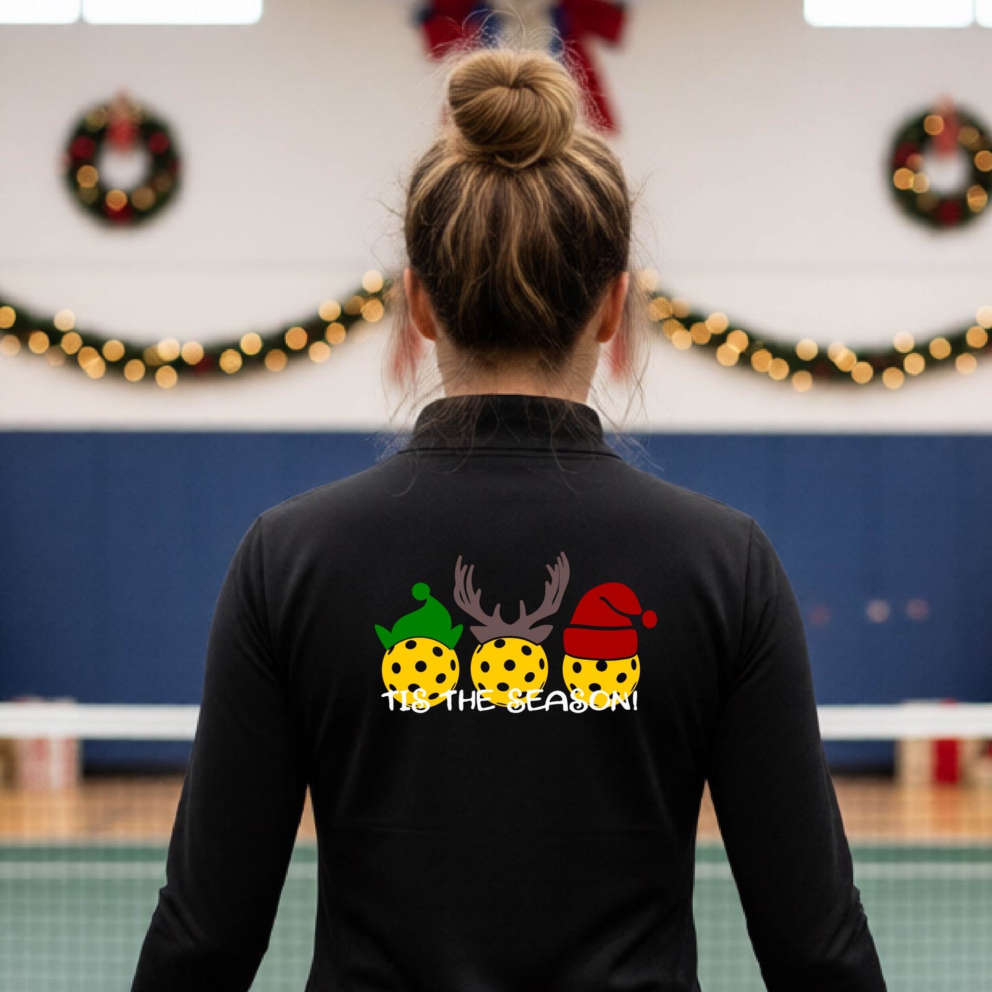 A woman viewed from behind wearing a black long-sleeve athletic top with a Christmas-themed graphic on the back. The graphic shows three yellow picKlebalLS with green, brown, and red holiday patterns, along with white text reading "TIS THE SEASON"